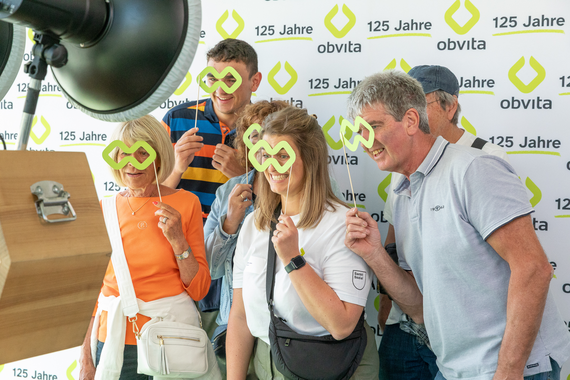 Eine Gruppe von fünf lächelnden Menschen steht dicht beieinander, trägt gelbe Pappbrillen und posiert spielerisch für ein Foto vor einem Hintergrund mit dem Schriftzug „obvita“. Zwei von ihnen halten eine hölzerne Brillen-Ausschnittfigur in der Hand.