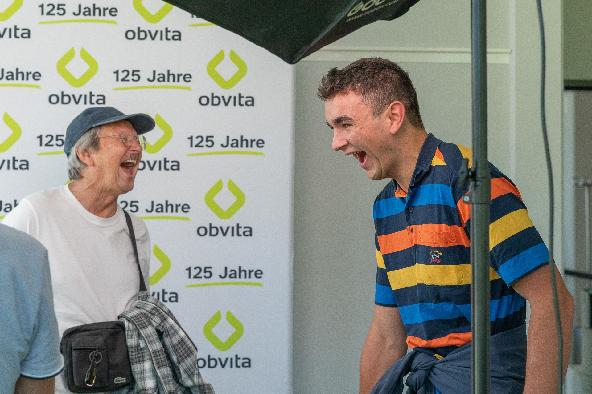 Zwei Männer lachen fröhlich beieinander bei einer Veranstaltung. Im Hintergrund ist wiederholt der Schriftzug „125 Jahre obvita“ zu sehen. Der eine Mann trägt eine blaue Mütze und eine Brille, der andere ein bunt gestreiftes Hemd.