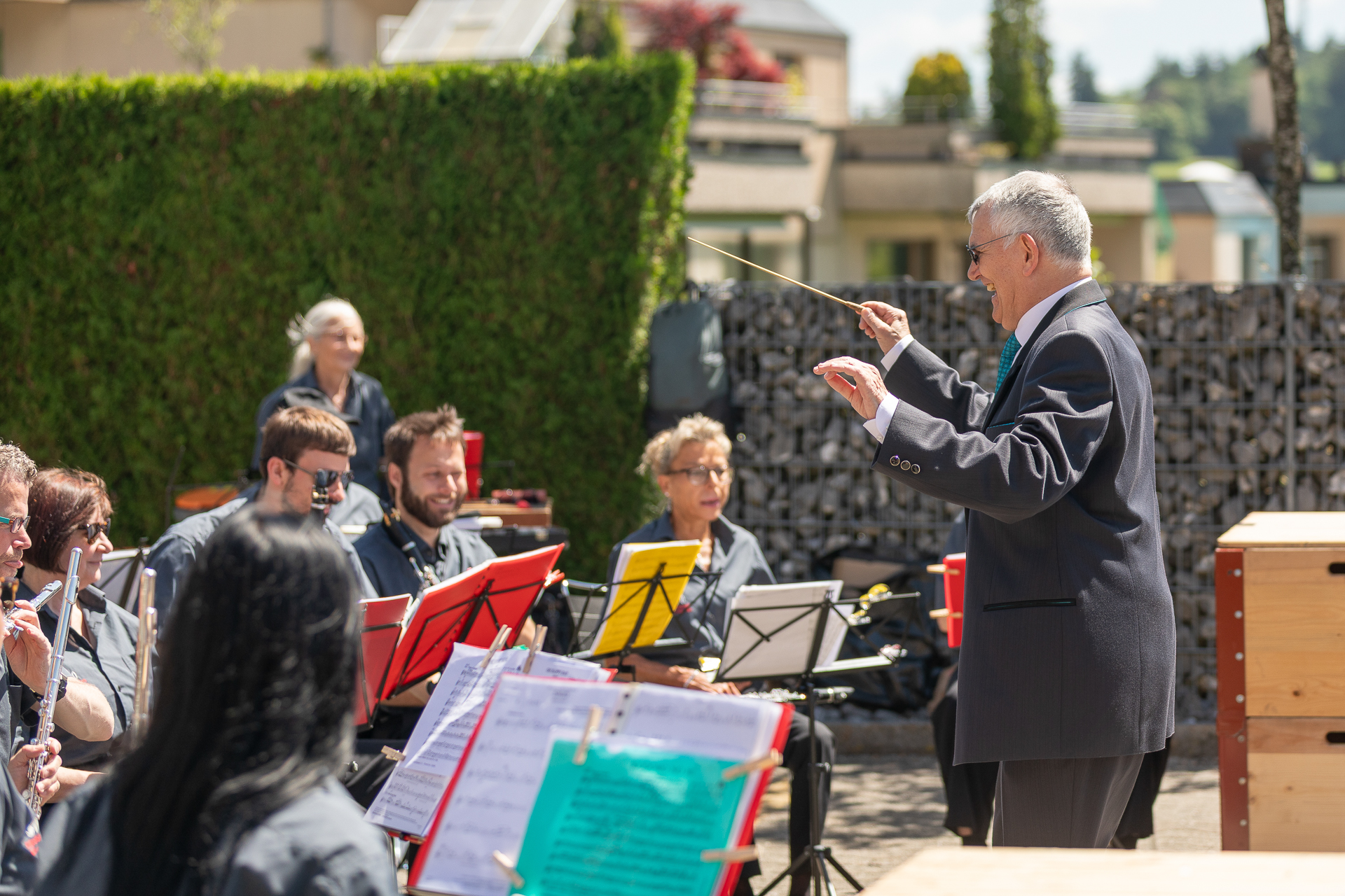 Ein Dirigent in einem dunklen Anzug leitet eine Orchesterprobe im Freien; die Musiker sitzen vor Notenständern und spielen unter strahlendem Sonnenschein vor einer grünen Kulisse.