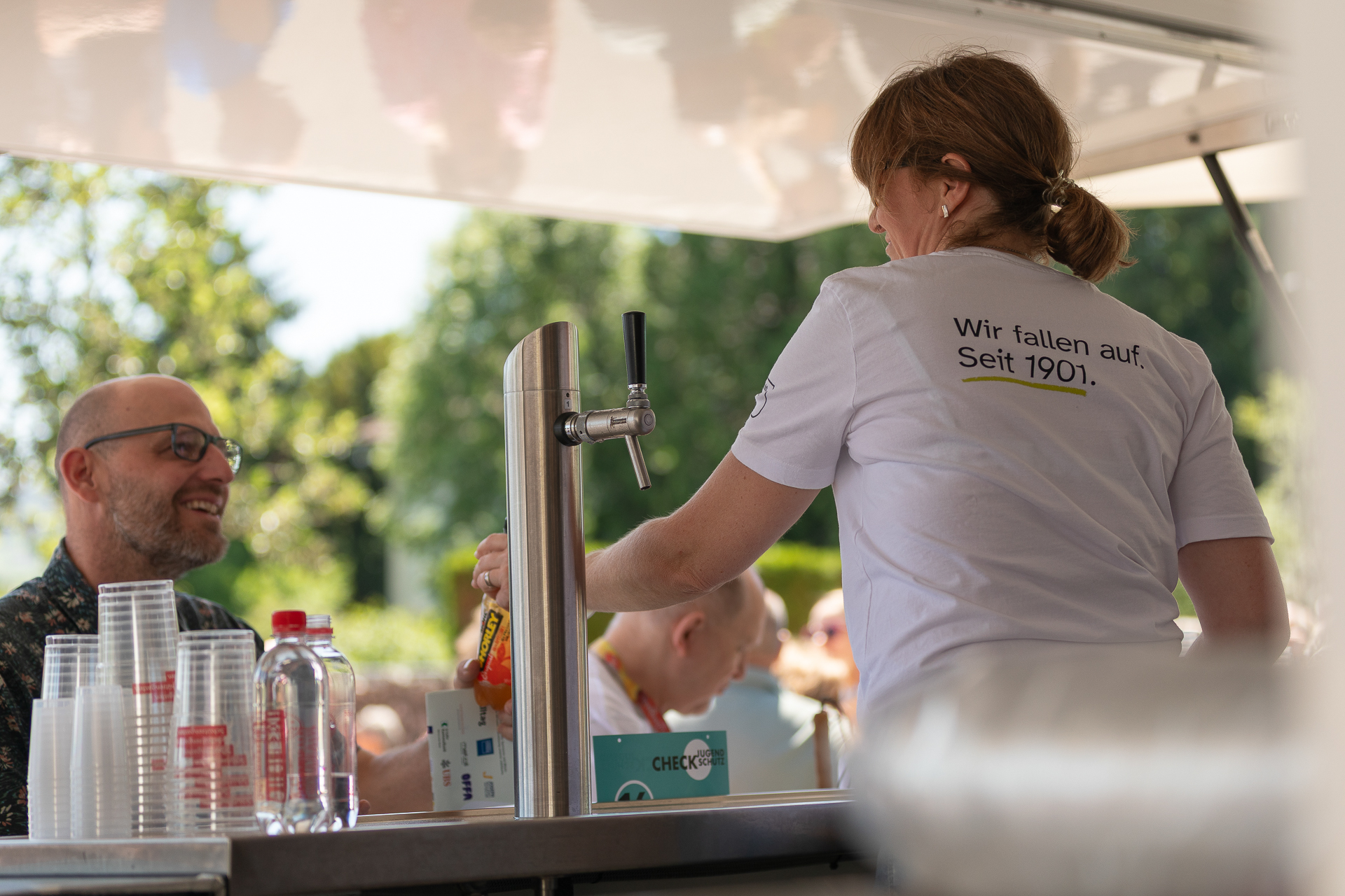 Eine Frau serviert an einer Theke im Freien Getränke und trägt ein weißes Shirt mit deutscher Aufschrift. Ein Mann sitzt ihr gegenüber und lächelt. Zu sehen sind Plastikbecher, Flaschen und ein Zapfhahn, im Hintergrund sind Pflanzen und Menschen zu erkennen.