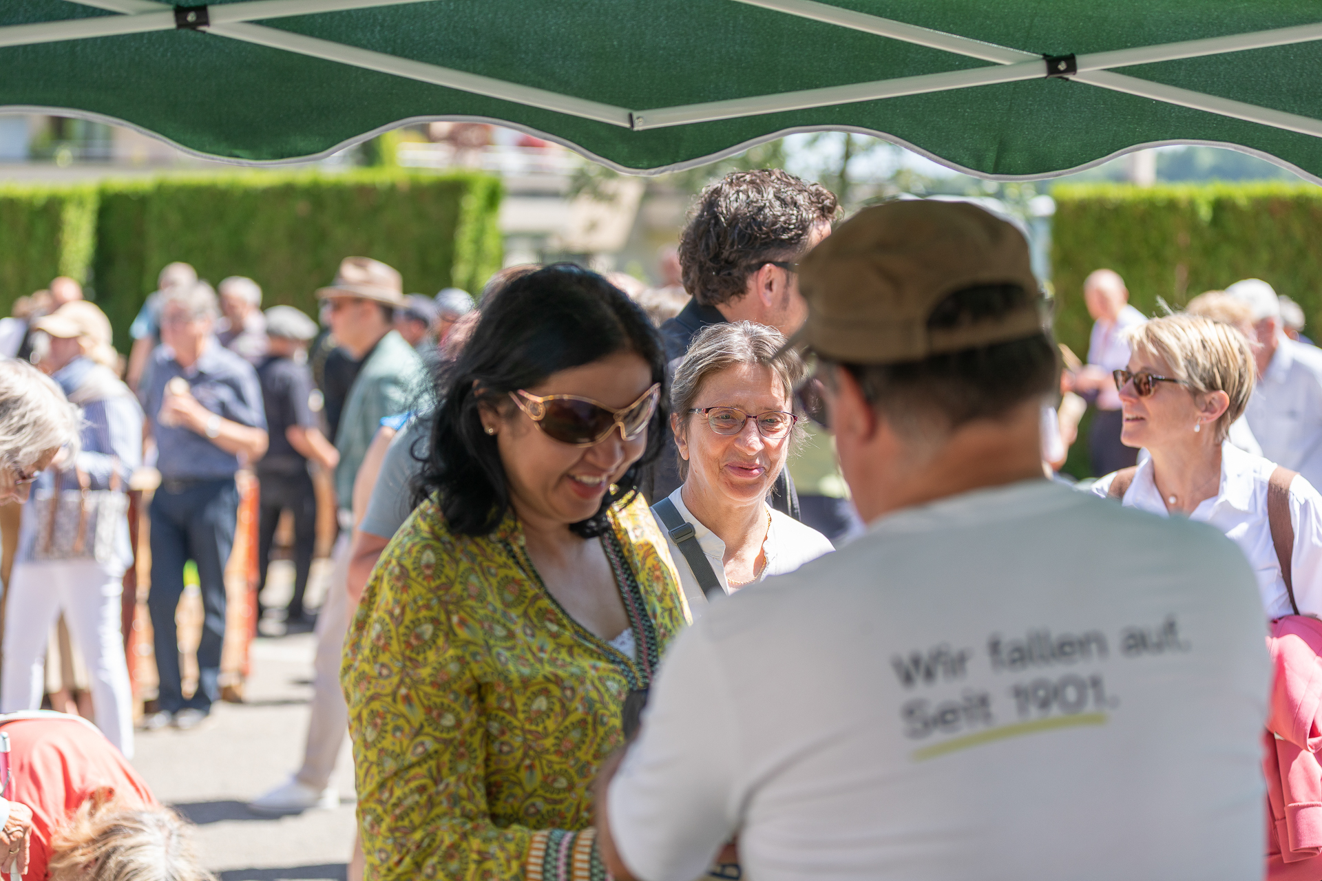 An einem sonnigen Tag trifft sich eine Gruppe von Menschen im Freien unter einem grünen Baldachin. Im Mittelpunkt stehen eine lächelnde Frau in gemusterter Kleidung und ein Mann in einem weißen Hemd mit einer Aufschrift auf dem Rücken. Im Hintergrund sind weitere Personen zu sehen.