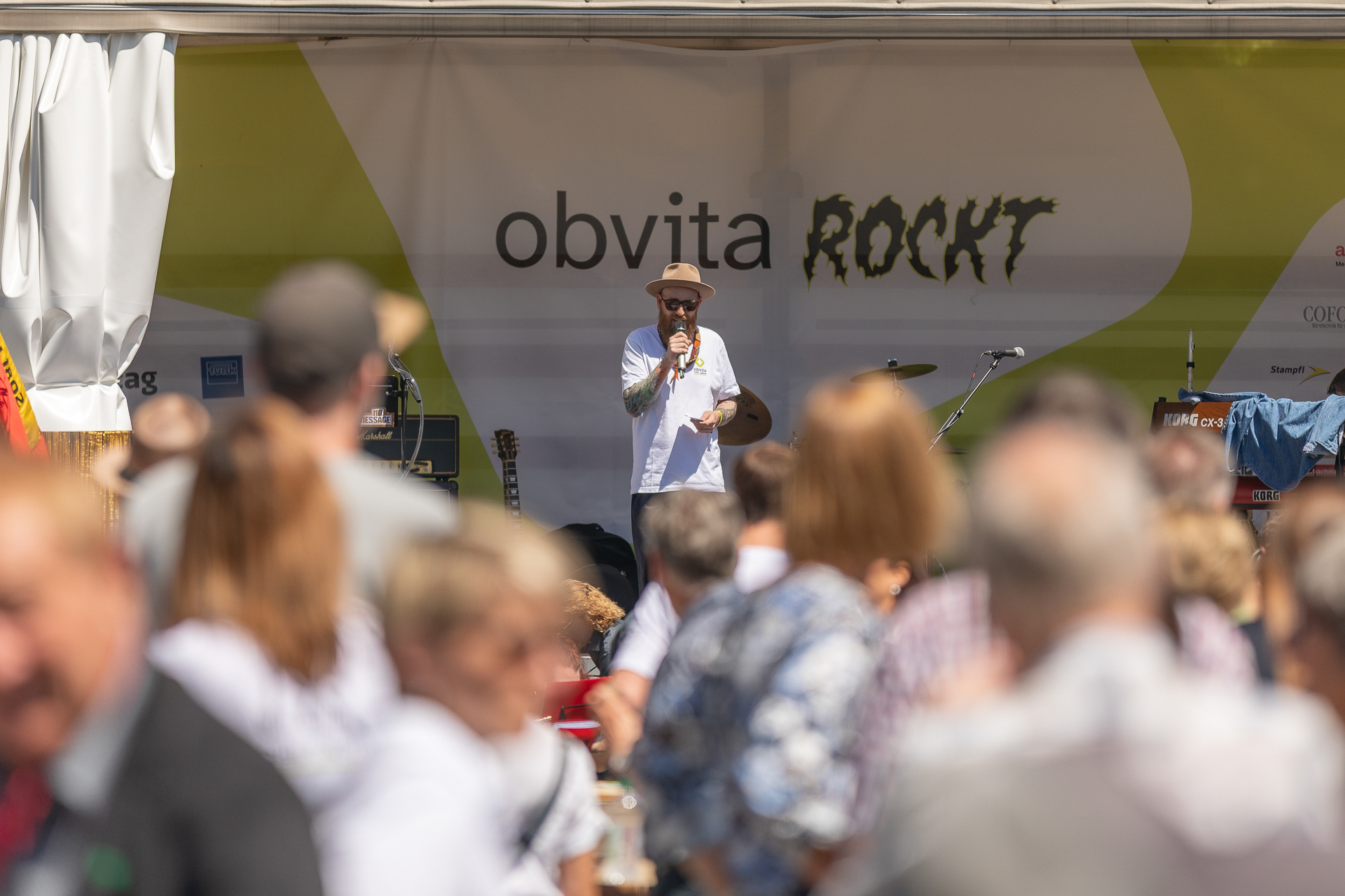 Ein Mann mit Hut und Sonnenbrille spricht auf einer mit „Obvita Rockt“ beschrifteten Außenbühne in ein Mikrofon und blickt dabei auf eine Menschenmenge bei einer sonnigen Veranstaltung.