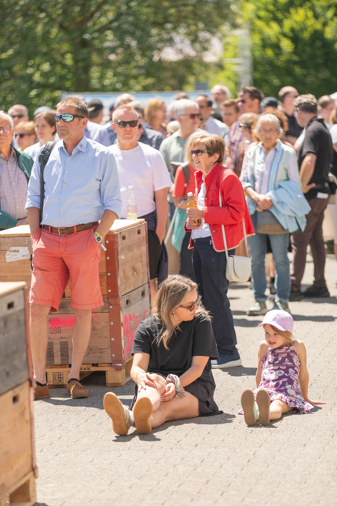 An einem sonnigen Tag steht eine Menschenmenge im Freien. Im Vordergrund sitzen eine Frau in Schwarz und ein junges Mädchen in einem lila Kleid und mit einem Hut auf dem Bürgersteig und lächeln. In der Nähe stehen Holzkisten. Im Hintergrund sind Bäume zu sehen.