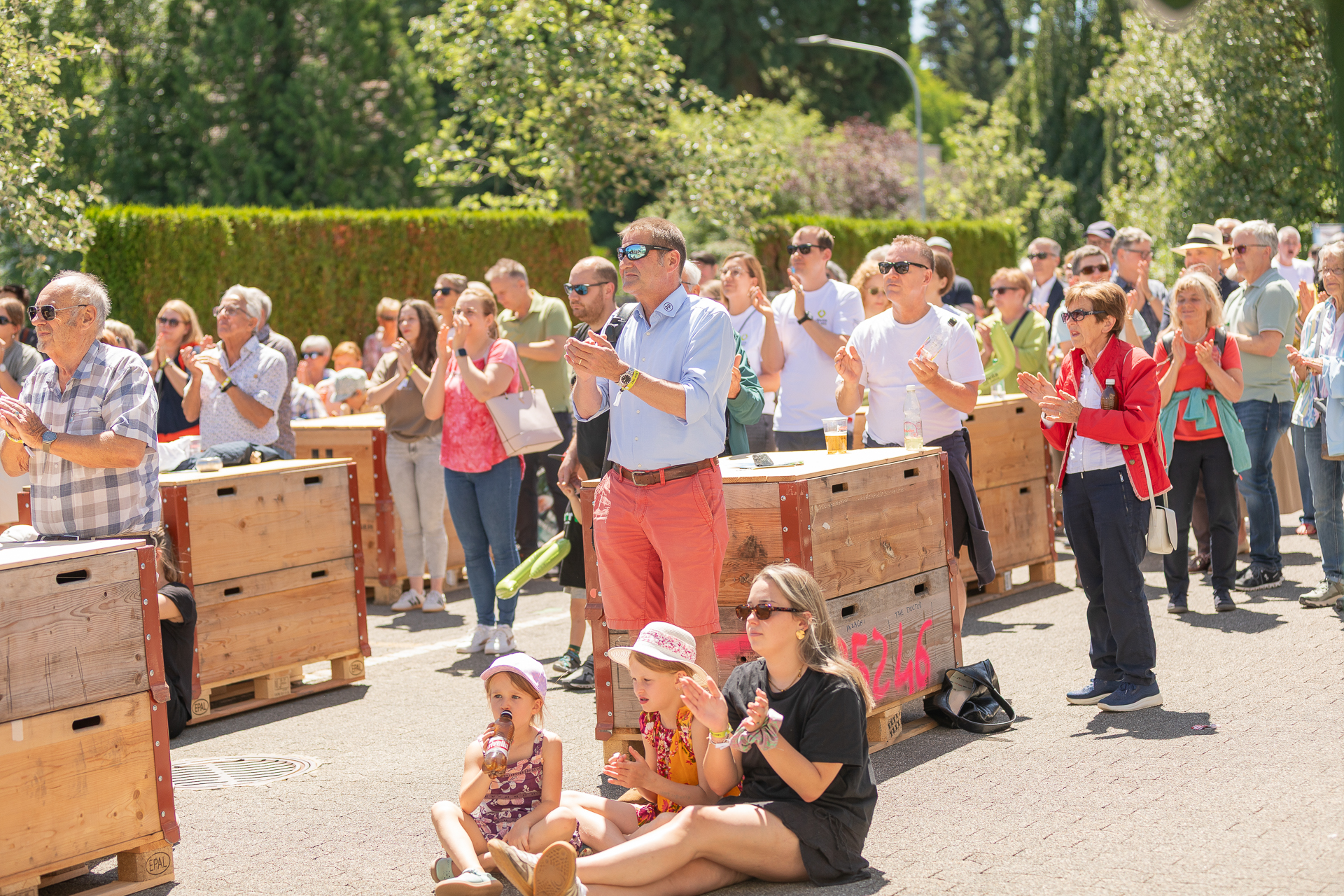 Eine bunte Menschenmenge, darunter Kinder und Erwachsene, steht und sitzt an einem sonnigen Tag im Freien, klatscht und verfolgt eine Veranstaltung. Einige sitzen auf dem Boden, während andere in der Nähe von Holzkisten stehen. Im Hintergrund sind Bäume und Grünflächen zu sehen.