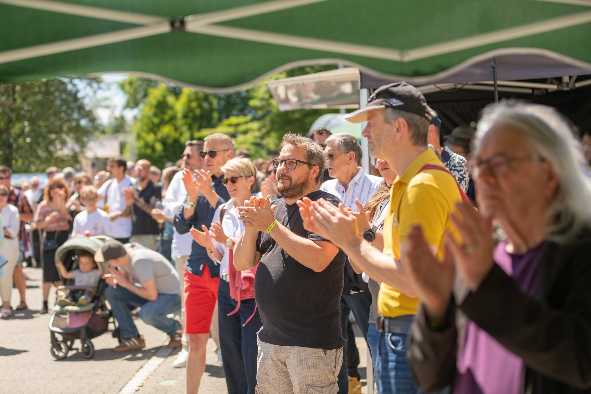 Eine bunte Menschenmenge steht im Freien unter einem grünen Vordach, klatscht und genießt an einem sonnigen Tag eine Veranstaltung. Einige tragen Sonnenbrillen, und im Hintergrund ist ein Kinderwagen mit einem Kind zu sehen.