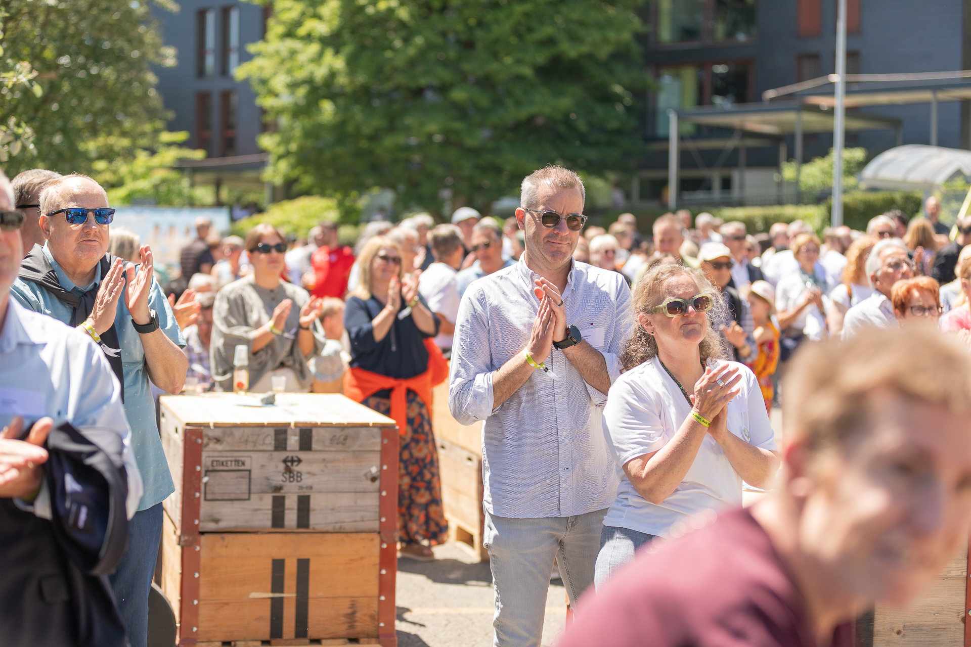 Eine große Gruppe von Menschen, von denen einige Sonnenbrillen tragen, steht an einem sonnigen Tag im Freien, klatscht in die Hände und beobachtet etwas, das sich außerhalb des Bildausschnitts befindet. Im Hintergrund sind Bäume, Holzkisten und moderne Gebäude zu sehen.