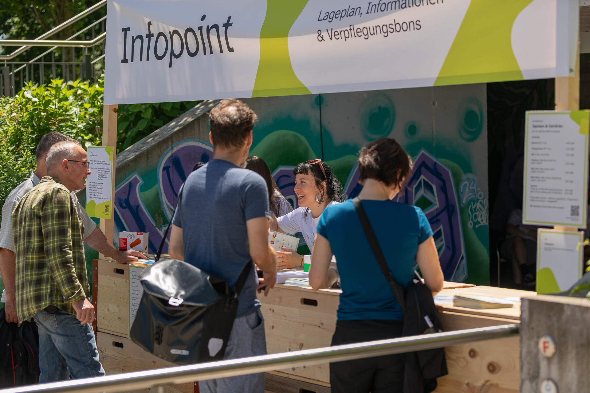 An einem Informationsstand im Freien mit der Aufschrift „Infopoint“ stehen mehrere Personen, denen ein lächelnder Mitarbeiter weiterhilft. Auf der Holztheke liegen Schilder und Broschüren. Im Hintergrund sind Grünpflanzen und eine Graffiti-Wand zu sehen.