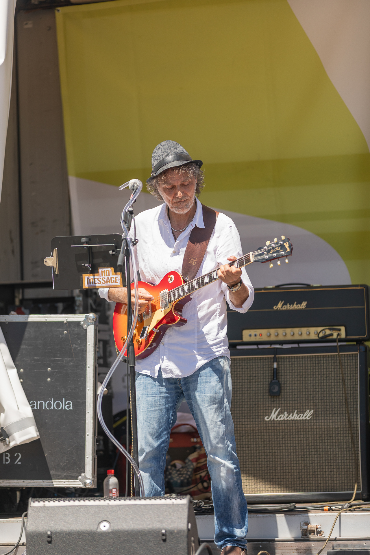 Ein Mann mit Hut und Brille spielt auf einer Außenbühne E-Gitarre; er steht vor Verstärkern und Musikgeräten, vor sich ein Mikrofon und ein Notenständer.
