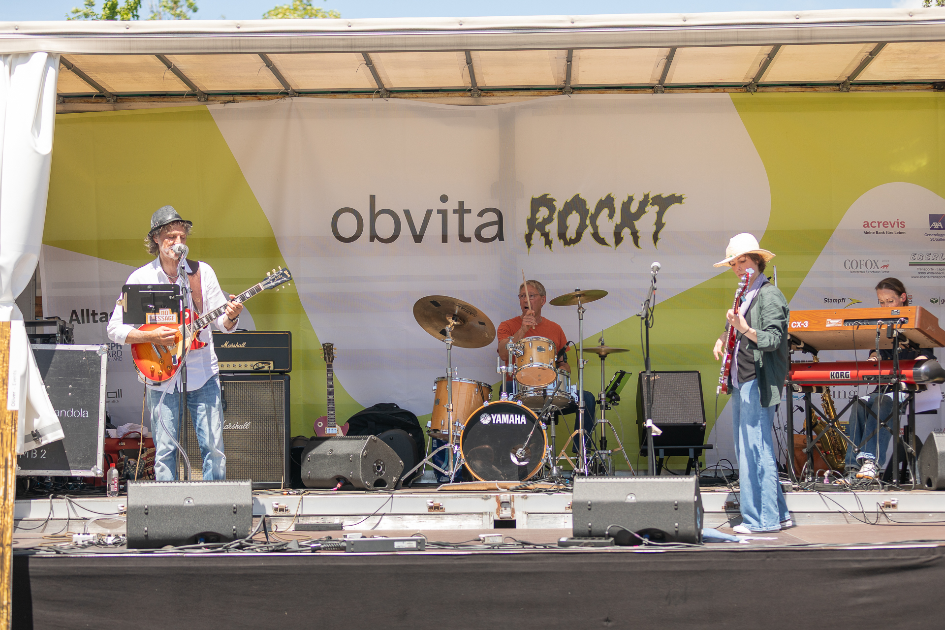 Eine Band tritt auf einer Open-Air-Bühne mit Gitarren, Schlagzeug, Keyboard und Mikrofonen auf. Auf einem Banner hinter ihnen steht „obvita ROCKT“. Vier Musiker spielen bei sonnigem Wetter.