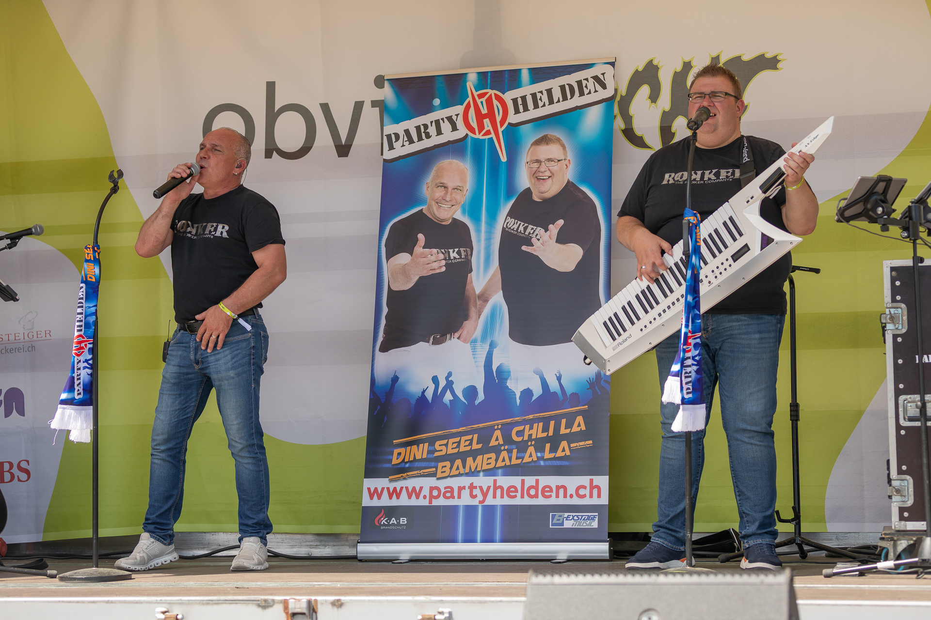 Zwei Männer treten auf der Bühne auf; einer singt ins Mikrofon, während der andere auf einer weißen Keytar spielt. Hinter ihnen ist ein leuchtendes Werbebanner mit ihren Fotos und der Aufschrift „Party Helden“ zu sehen. Mikrofone und Lautsprecher sind zu erkennen.