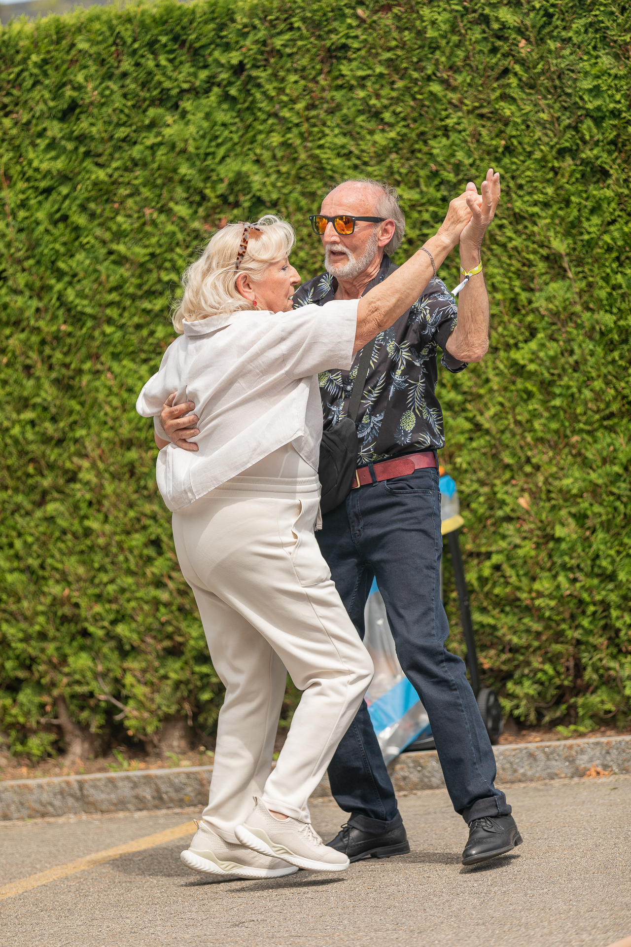 Ein älteres Ehepaar tanzt an einem sonnigen Tag fröhlich im Freien und lächelt sich dabei an. Die beiden sind leger gekleidet, im Hintergrund sind Grünflächen und eine hohe Hecke zu sehen.