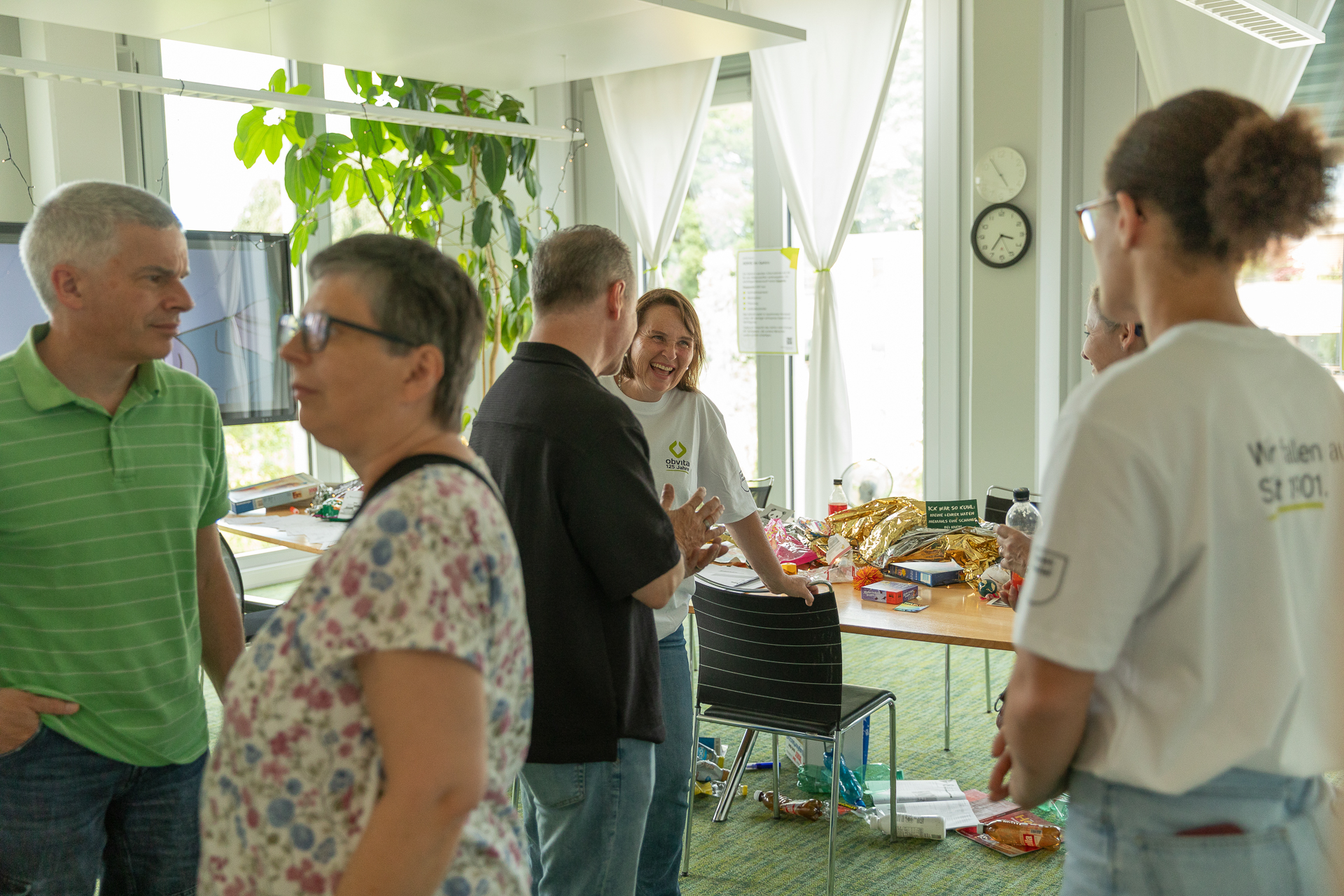 In einem hellen Raum mit großen Fenstern und Pflanzen unterhält sich eine Gruppe von Menschen und lacht. Im Hintergrund steht ein Tisch mit Snacks und Getränken. Einige tragen weiße Hemden, und eine Wanduhr ist zu sehen.