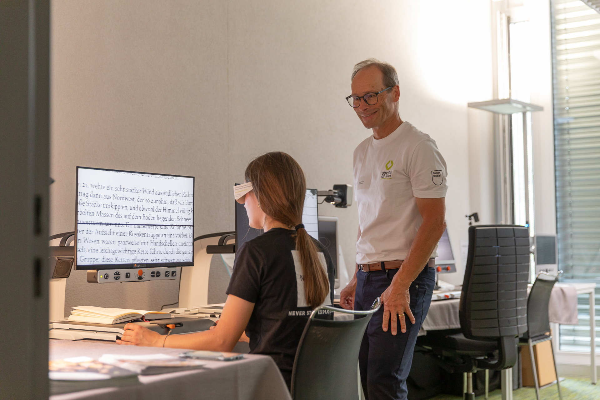 Eine junge Frau nutzt einen großen Bildschirm mit vergrößertem Text, während ein älterer Mann lächelnd neben ihr steht. Beide befinden sich in einem modernen, gut beleuchteten Büro mit Computern und Stühlen.