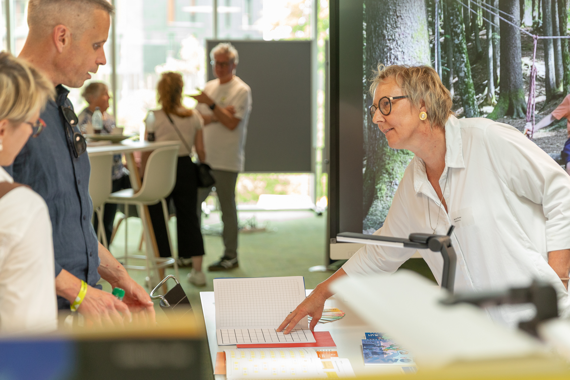 Eine Frau mit Brille und weißem Hemd deutet auf ein offenes Notizbuch auf einem Tisch, während sie bei einer Veranstaltung oder Ausstellung in einem Saal mit zwei Personen spricht. Im Hintergrund sind weitere Personen und Ausstellungsstücke zu sehen.