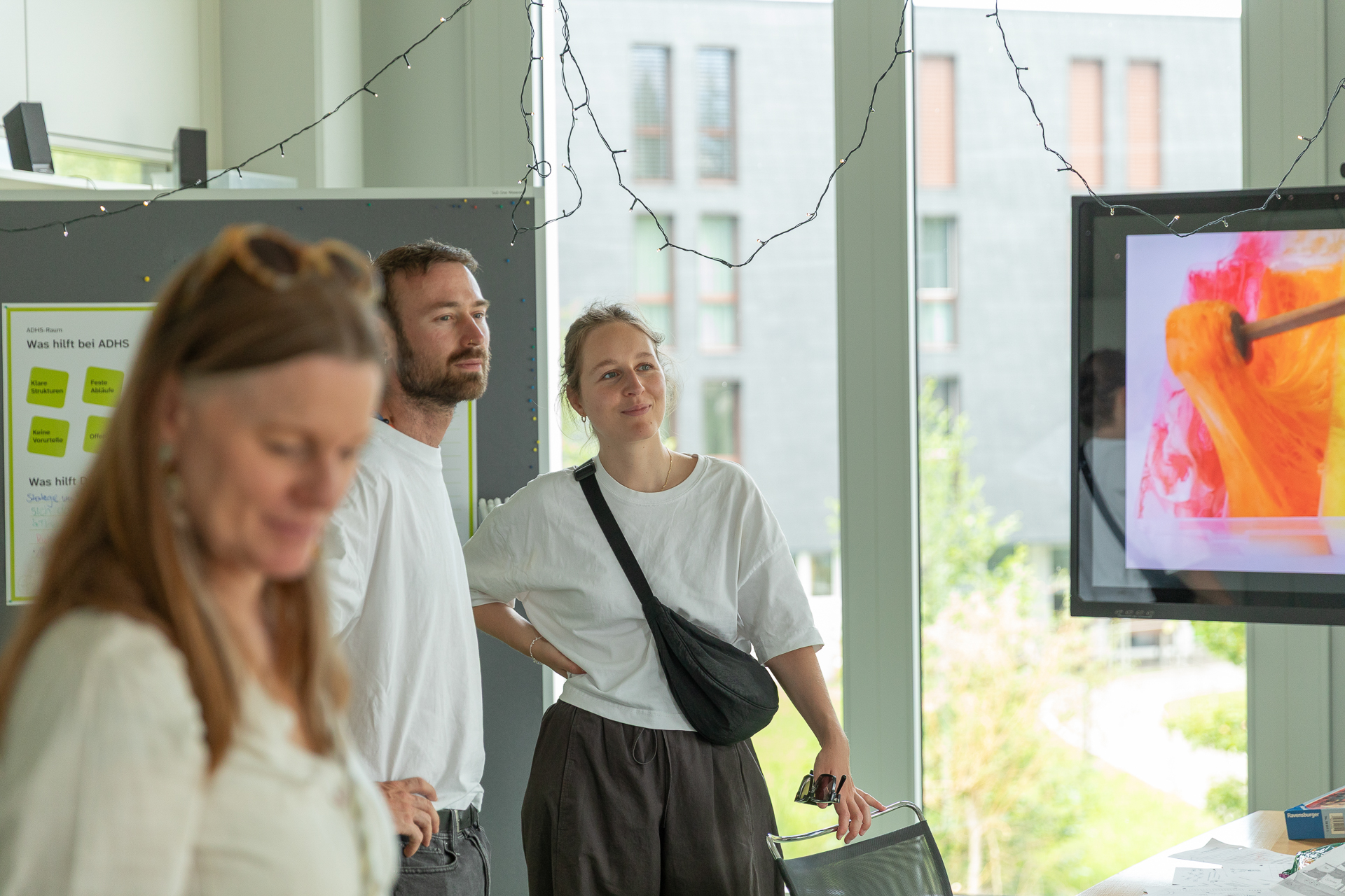 Drei Personen stehen in einem hellen Raum neben einem Bildschirm, auf dem farbenfrohe abstrakte Kunst zu sehen ist. Zwei von ihnen, ein Mann und eine Frau, betrachten den Bildschirm aufmerksam; eine dritte Frau ist im Vordergrund nur verschwommen zu erkennen.