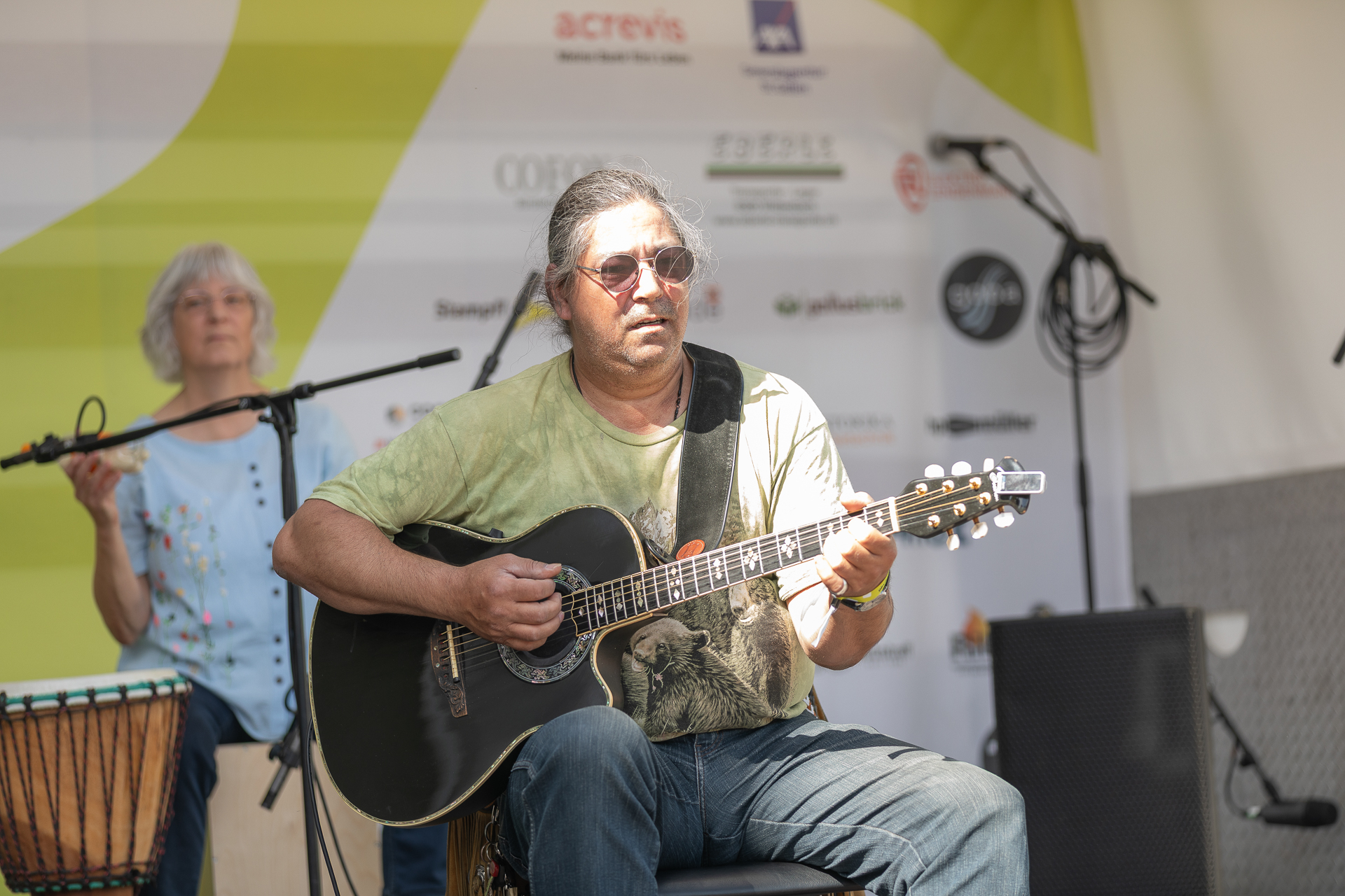 Ein Mann mit langen grauen Haaren und Sonnenbrille spielt auf der Bühne Akustikgitarre, während eine Frau im Hintergrund die Trommel spielt. Hinter ihnen ist eine Kulisse mit verschiedenen Sponsorenlogos zu sehen.
