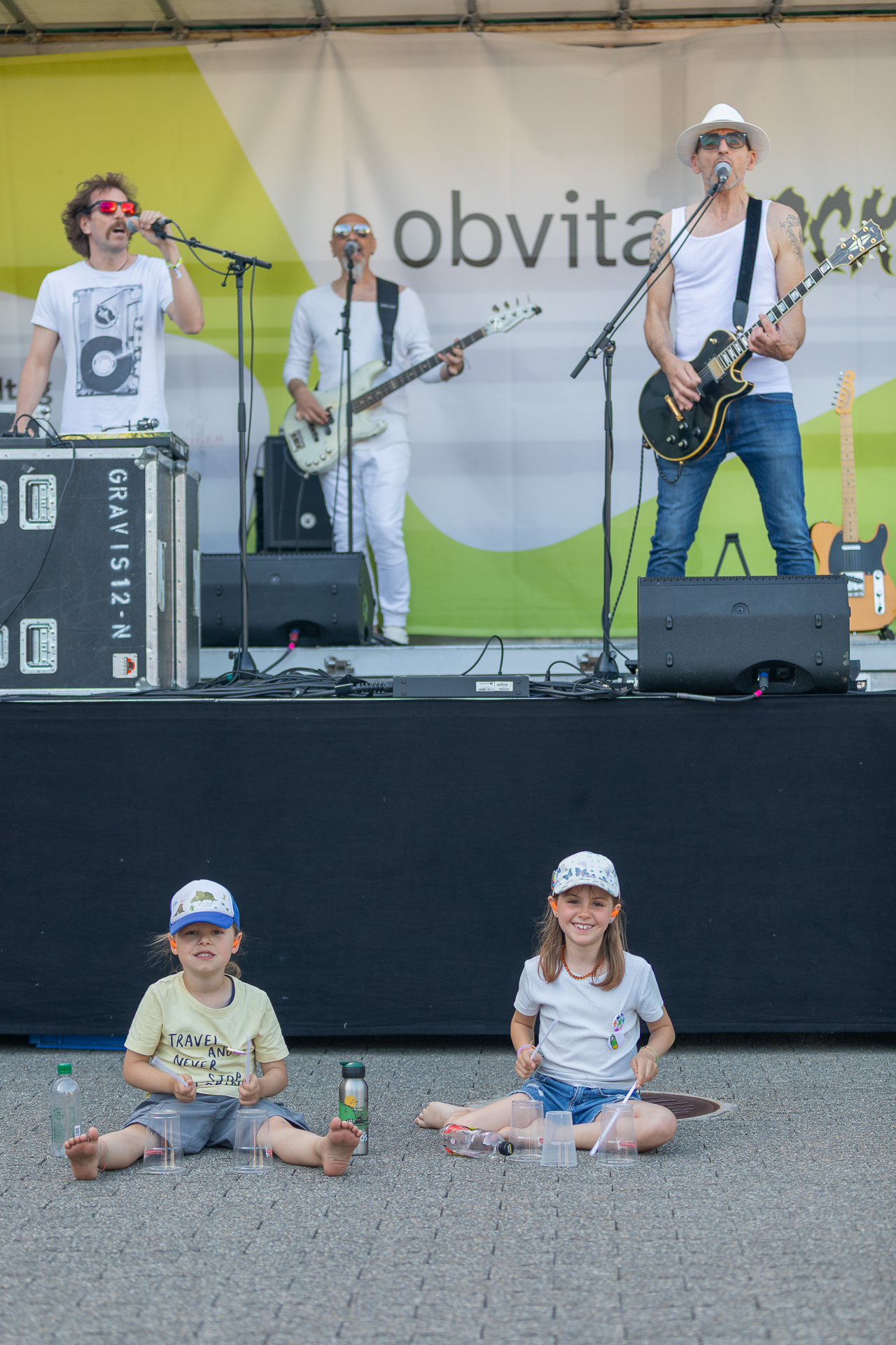 Zwei Kinder sitzen auf Matten vor einer Freiluftbühne, lächeln und halten Getränke in den Händen. Hinter ihnen spielt eine vierköpfige Band – bestehend aus zwei Gitarristen, einem Bassisten und einem Sänger – unter einem grün-weißen Banner.