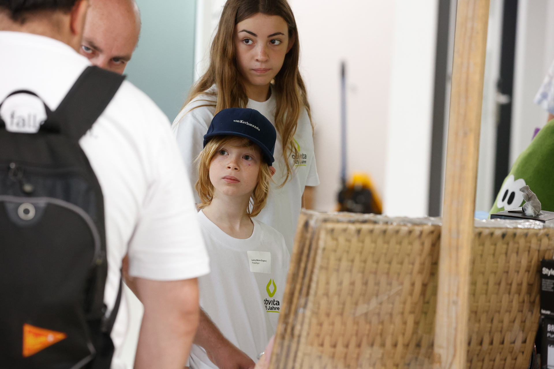 Ein junges Mädchen und ein Kind mit einer blauen Mütze, beide in weißen Hemden, schauen einem Mann mit Rucksack in einem Raum aufmerksam zu. Sie scheinen an einer Veranstaltung oder einem Workshop teilzunehmen.