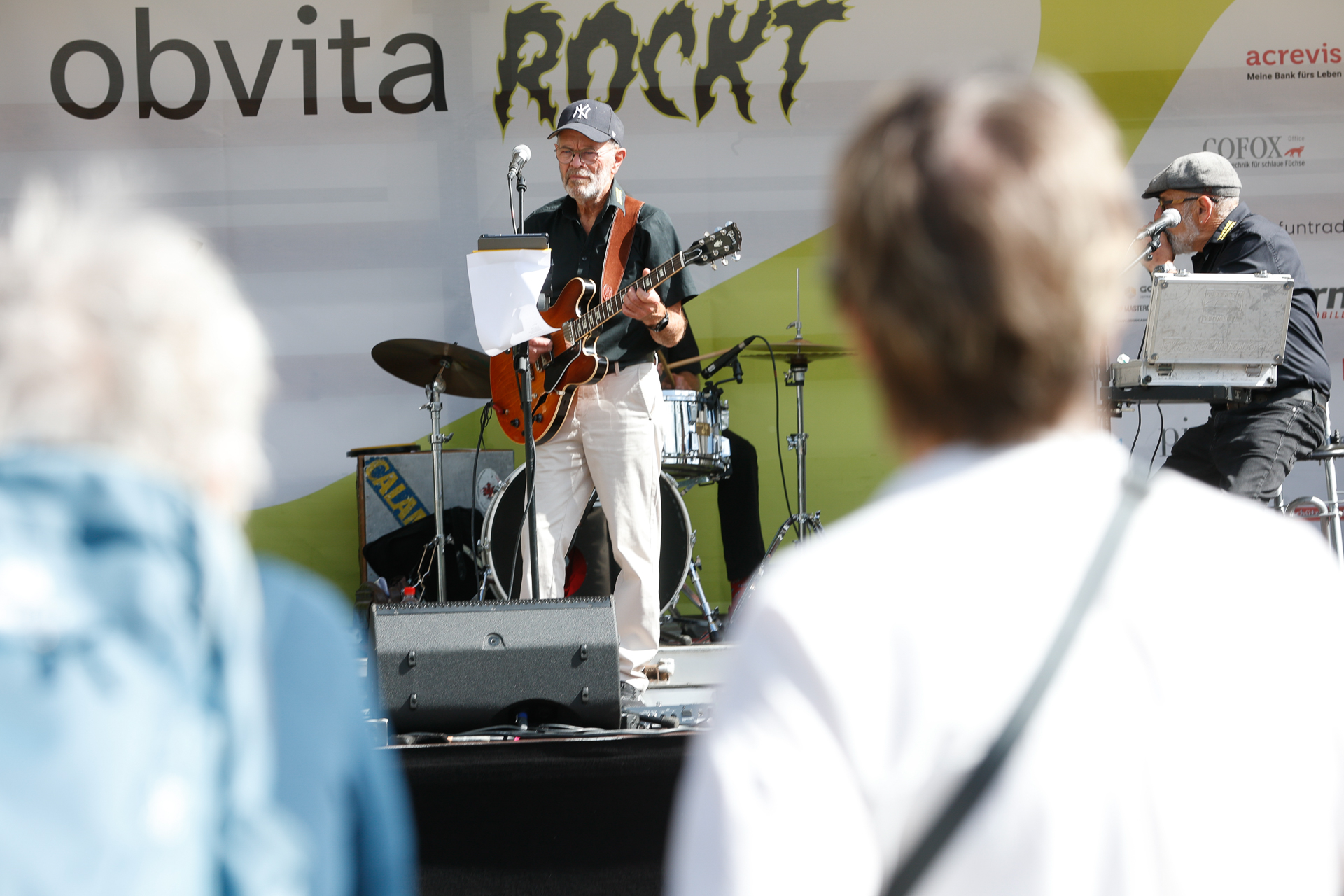 Ein Gitarrist und Sänger tritt mit einer Band auf einer Open-Air-Bühne auf, während das Publikum zusieht. Auf dem Bühnenhintergrund steht in großen Buchstaben „obvita ROCKT“.