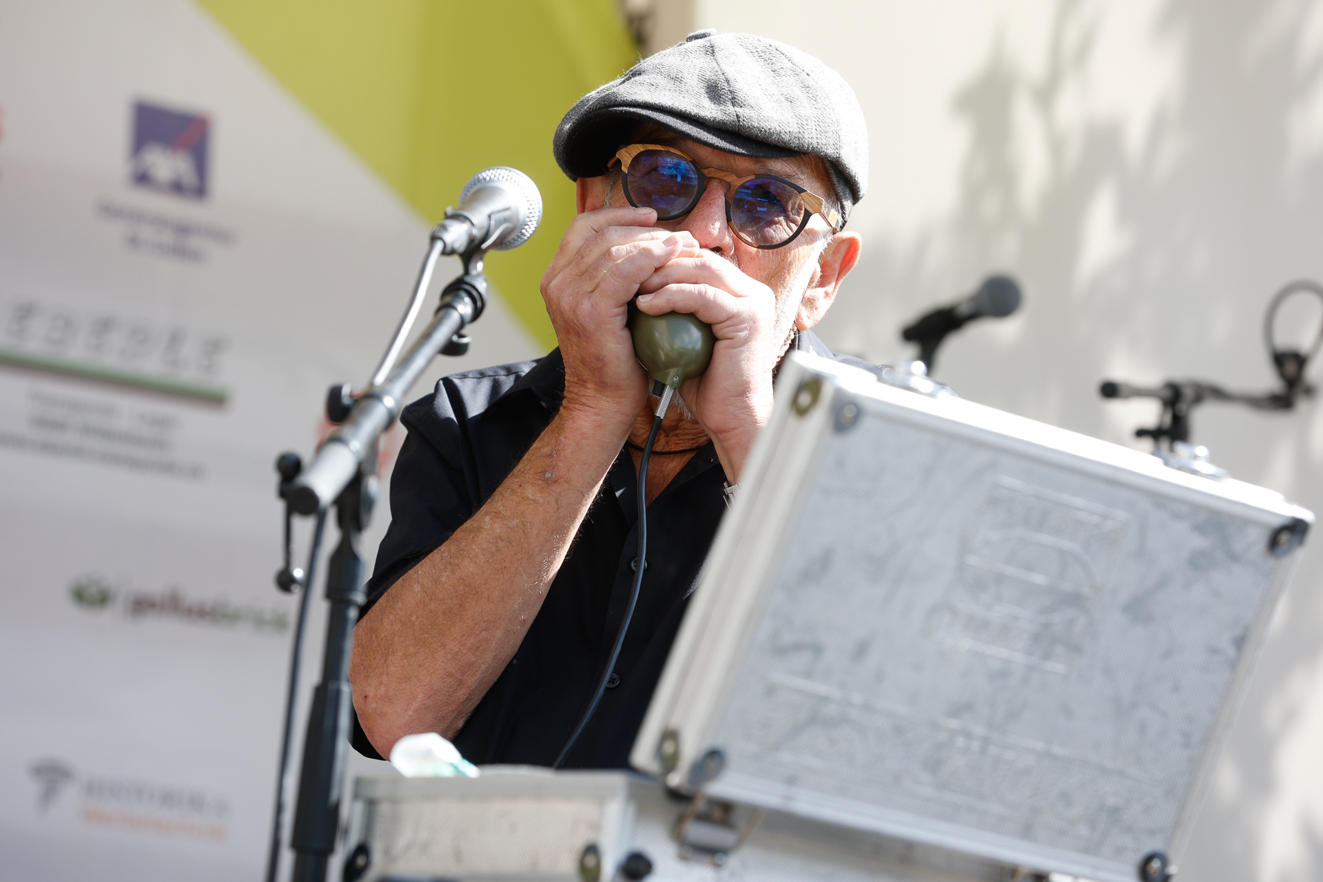 Ein Mann mit Sonnenbrille und Schirmmütze spielt bei einer Veranstaltung im Freien hinter einem Metallkoffer stehend Mundharmonika in ein Mikrofon.