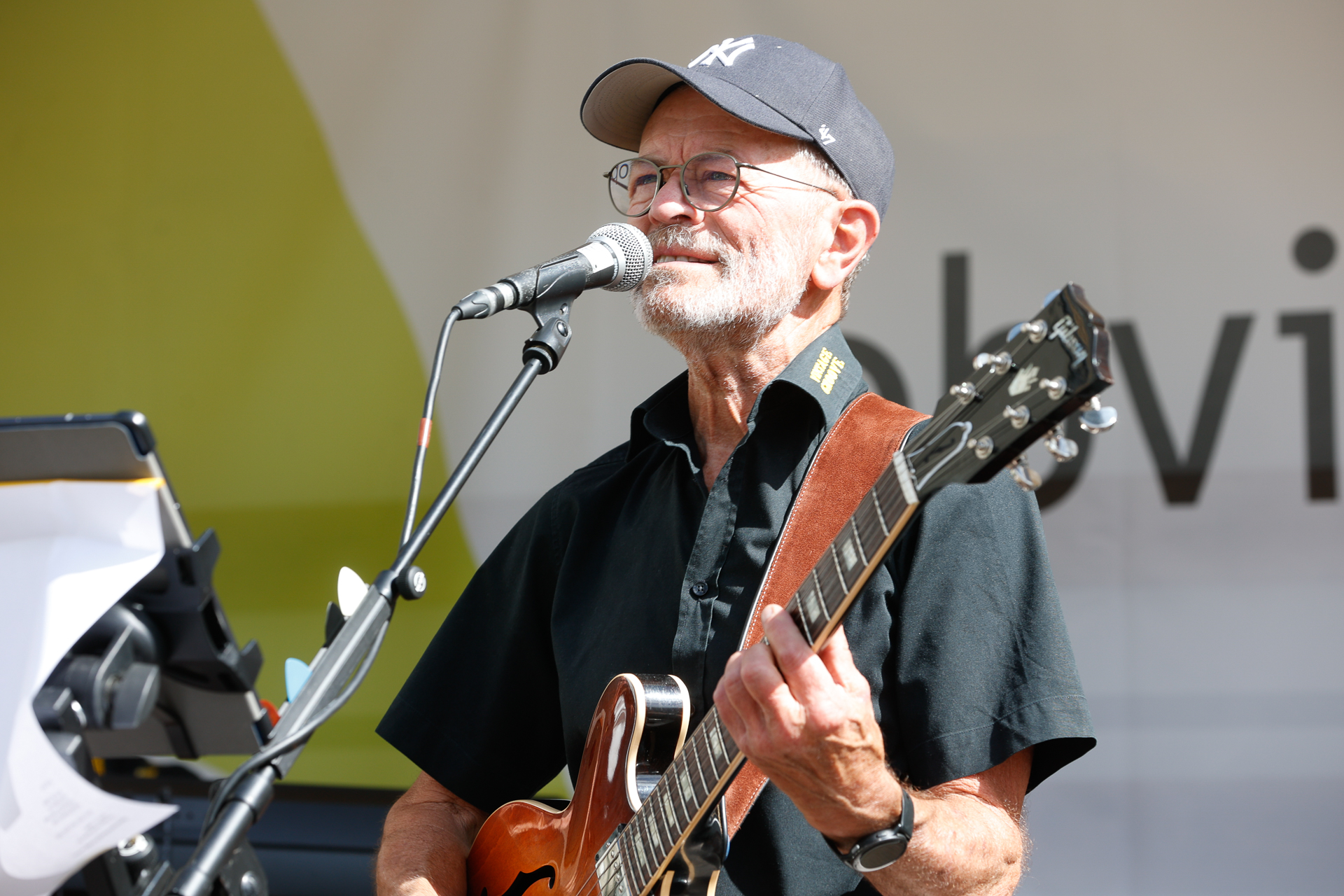 Ein älterer Mann mit Brille und Baseballmütze spielt auf einer Außenbühne E-Gitarre und singt ins Mikrofon. Er trägt ein dunkles Hemd mit kurzen Ärmeln.