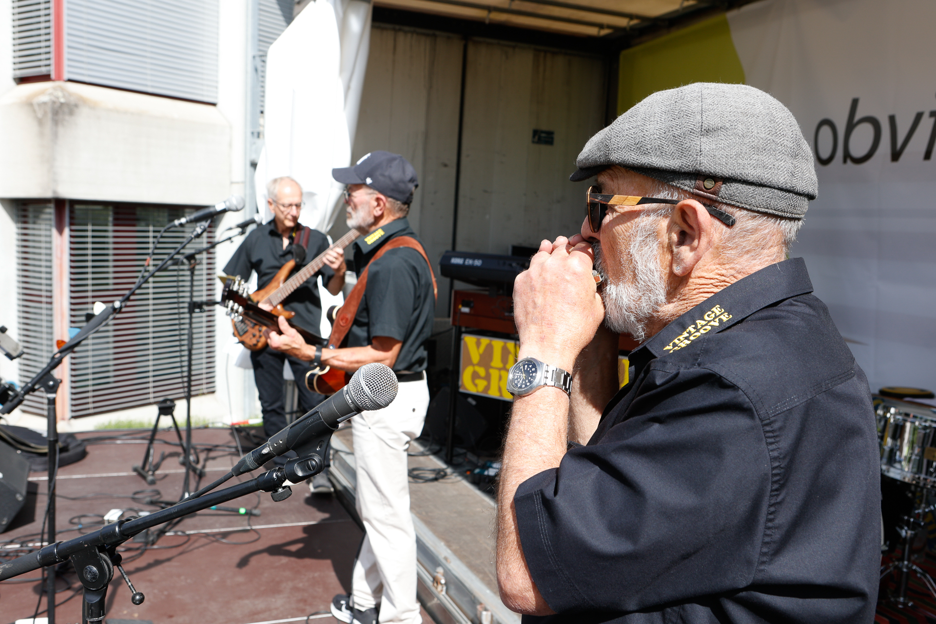 Drei ältere Männer treten auf einer Freiluftbühne auf; einer spielt Gitarre, ein anderer Bass und der dritte Mundharmonika. Alle tragen schwarze Hemden und Hüte. Um sie herum sind Mikrofone und Musikinstrumente zu sehen.