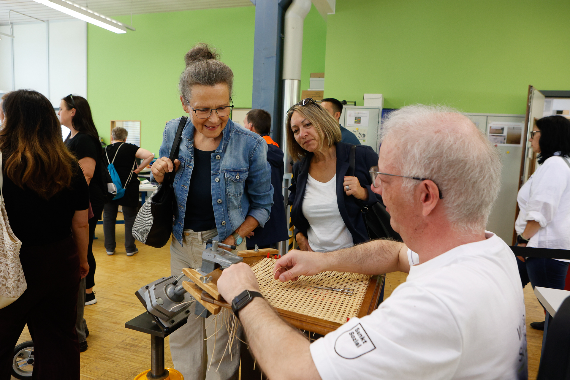 Ein Mann führt das Weben an einem Webstuhl vor, während zwei Frauen ihm zusehen und lächeln. Die Szene spielt in einem belebten Innenraum mit grünen Wänden und mehreren Personen im Hintergrund.