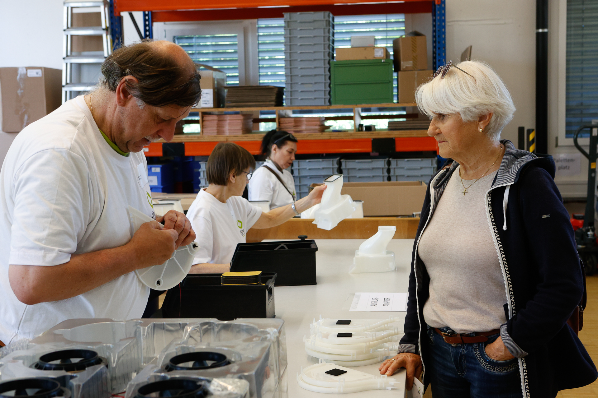 Ein Mann und eine ältere Frau stehen an einem Tisch und montieren Kunststoffteile, während zwei weitere Frauen im Hintergrund in einem gut beleuchteten Industriebereich mit Regalen und Kisten arbeiten.
