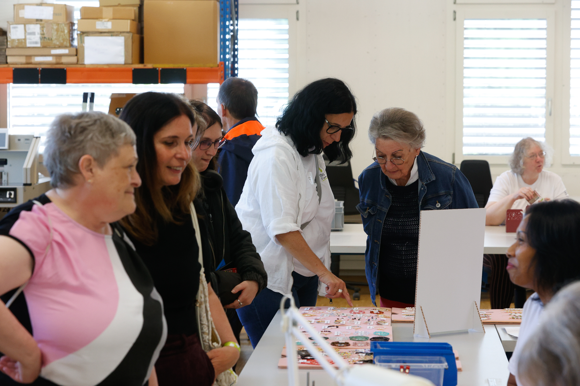 Eine Gruppe von Frauen sitzt drinnen um einen Tisch herum und betrachtet Gegenstände, die zum Verkauf oder zur Ausstellung ausgestellt sind. Einige unterhalten sich und lächeln; es scheint sich um eine Gemeinschaftsveranstaltung oder einen Workshop zu handeln. Im Hintergrund sind Regale und Kisten zu sehen.