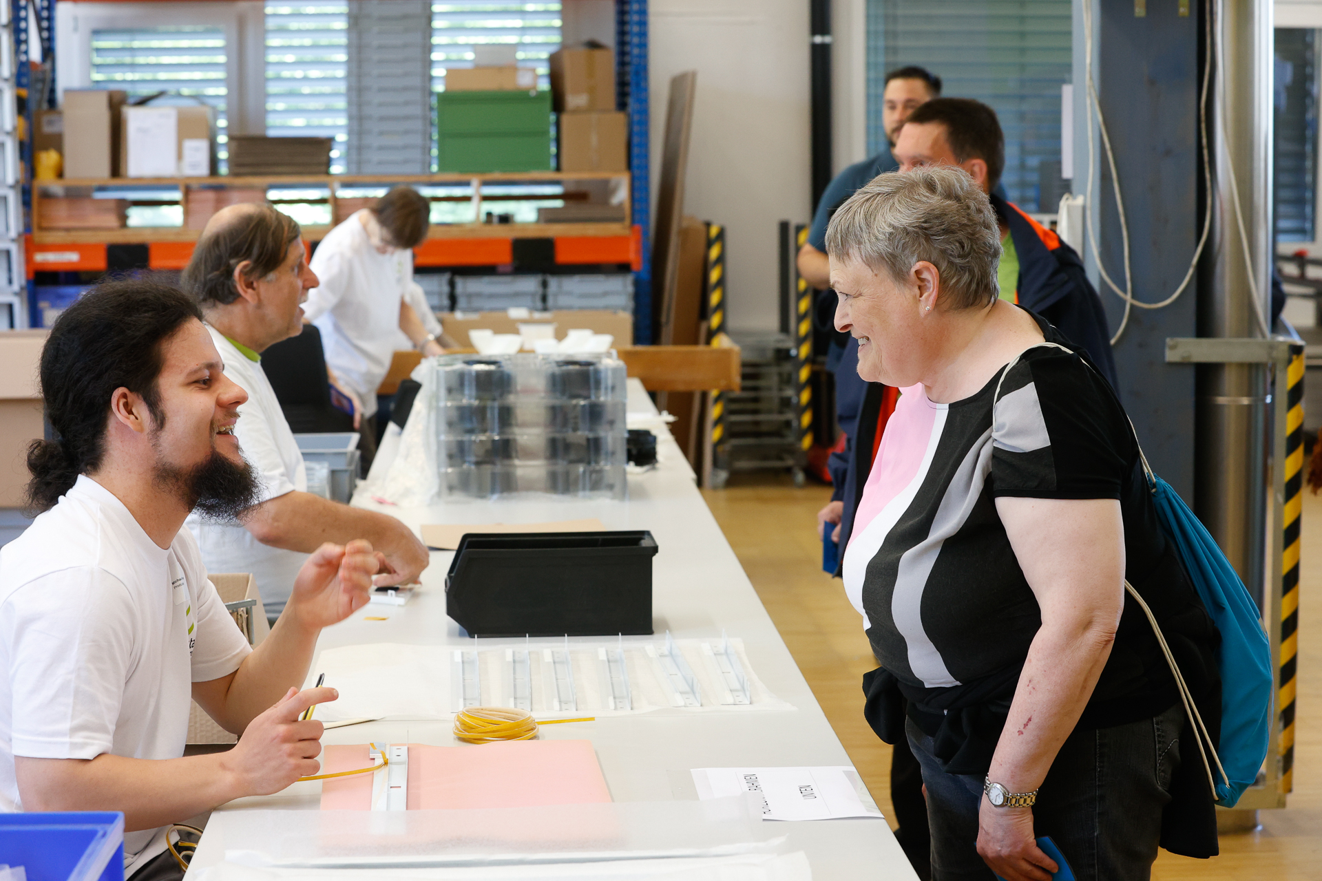 Ein Mann, der an einem Tisch sitzt, lächelt und unterhält sich mit einer älteren Frau, die ihm in einem hellen Raum gegenüber steht; im Hintergrund sind Menschen bei der Arbeit zu sehen und Materialien, die ordentlich in Regalen aufgereiht sind.