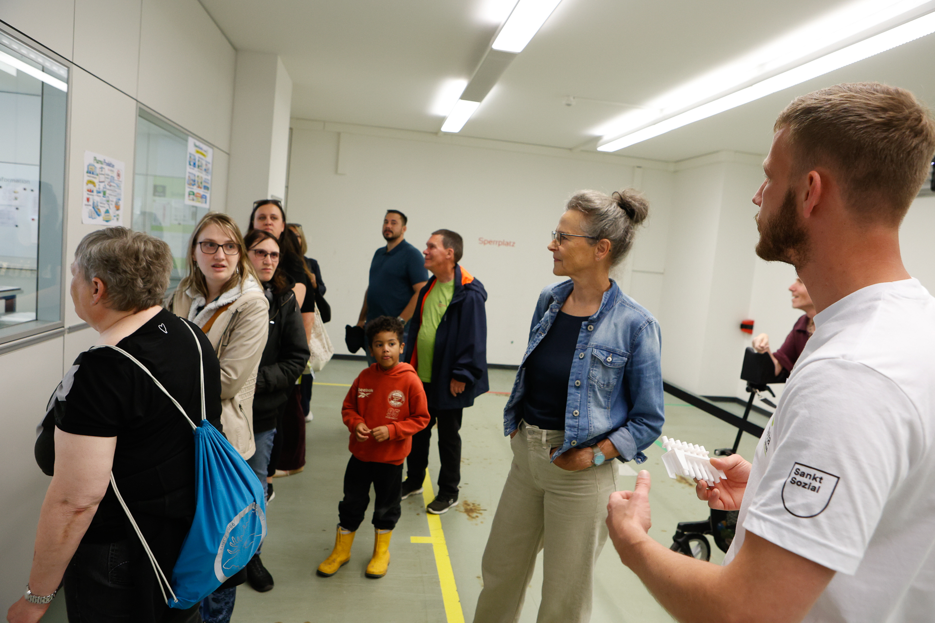 Eine Gruppe aus Erwachsenen und Kindern steht in einem Flur; einige betrachten Zettel an der Wand, während ein Mann in einem weißen Hemd einen kleinen Gegenstand in der Hand hält und unter grellem Neonlicht mit ihnen spricht.
