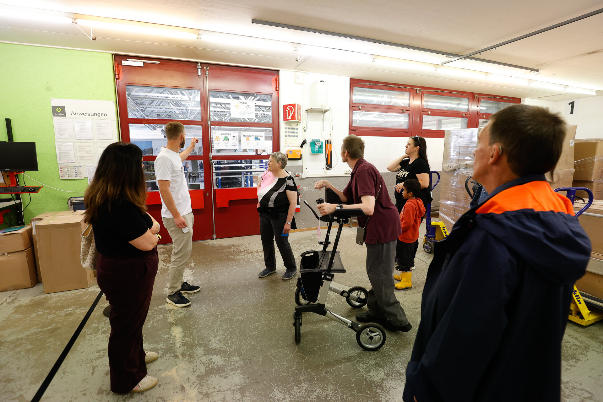 Eine Gruppe von Menschen, darunter eine Person mit einem Rollator, steht im Inneren einer Lagerhalle und unterhält sich, während im Hintergrund rote Türen, Kartons und Hubwagen zu sehen sind.