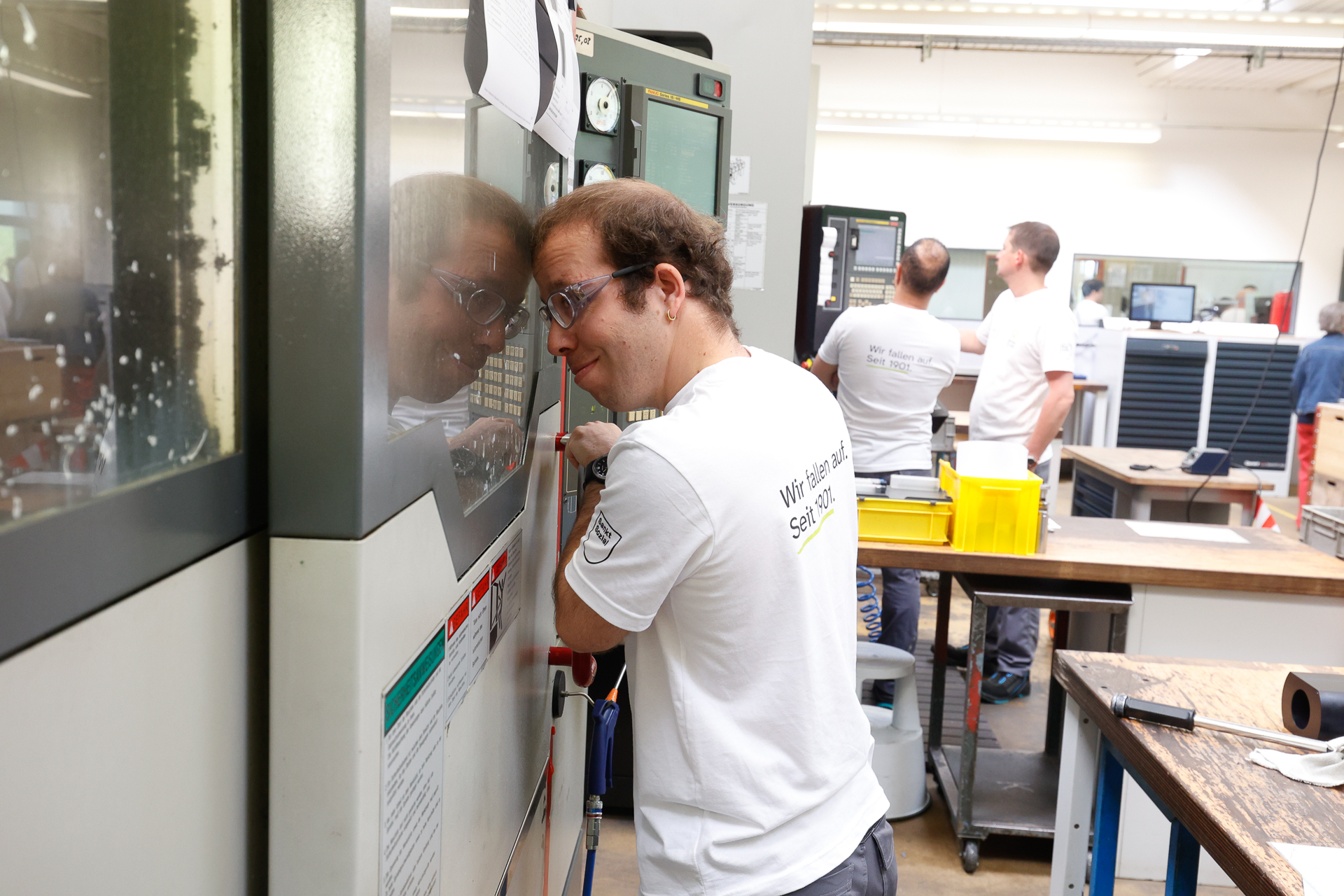 Ein Mann mit Schutzbrille bedient eine Maschine in einer hell beleuchteten Werkstatt, während im Hintergrund zwei weitere Personen arbeiten. Er konzentriert sich auf das Bedienfeld, und sein Spiegelbild ist auf der Oberfläche der Maschine zu sehen.