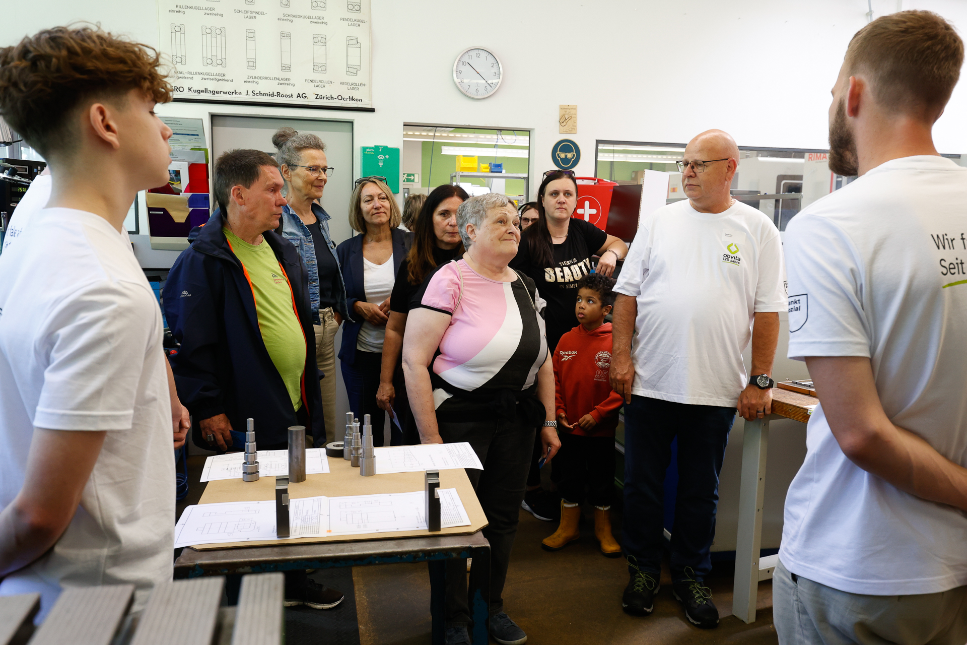 Eine Gruppe von Menschen, darunter Erwachsene und Kinder, steht in einem Workshop oder Klassenzimmer und hört zwei Kursleitern in weißen Hemden zu. Im Hintergrund sind Maschinen, Plakate und eine Uhr zu sehen.