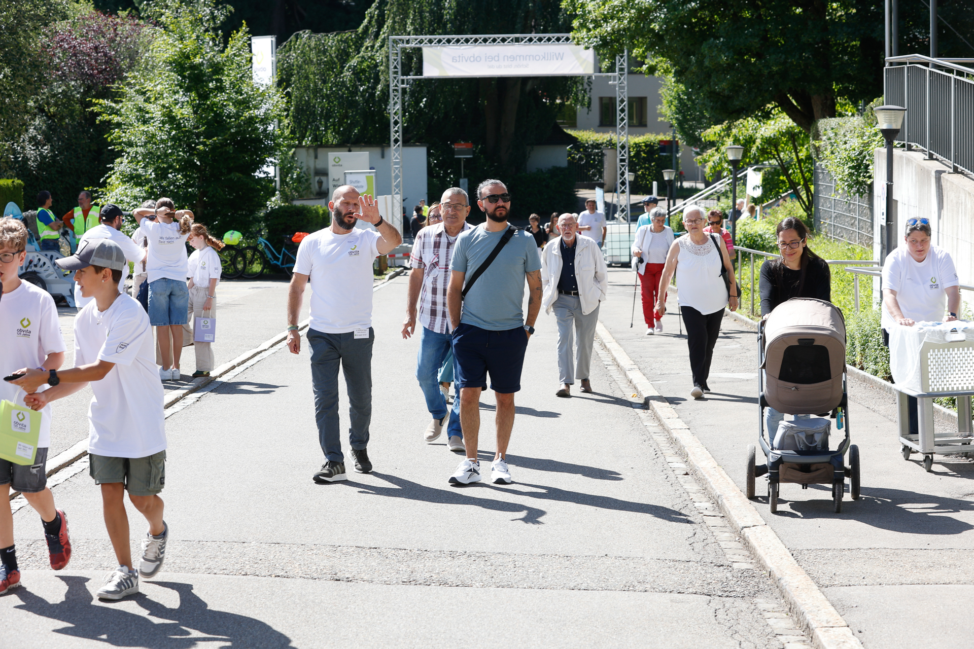 Eine Gruppe von Menschen unterschiedlichen Alters spaziert an einem sonnigen Tag im Freien; einige tragen weiße Hemden, eine Person schiebt einen Kinderwagen, und andere gehen gemeinsam auf einem gepflasterten Weg spazieren, der von Grünflächen gesäumt ist.