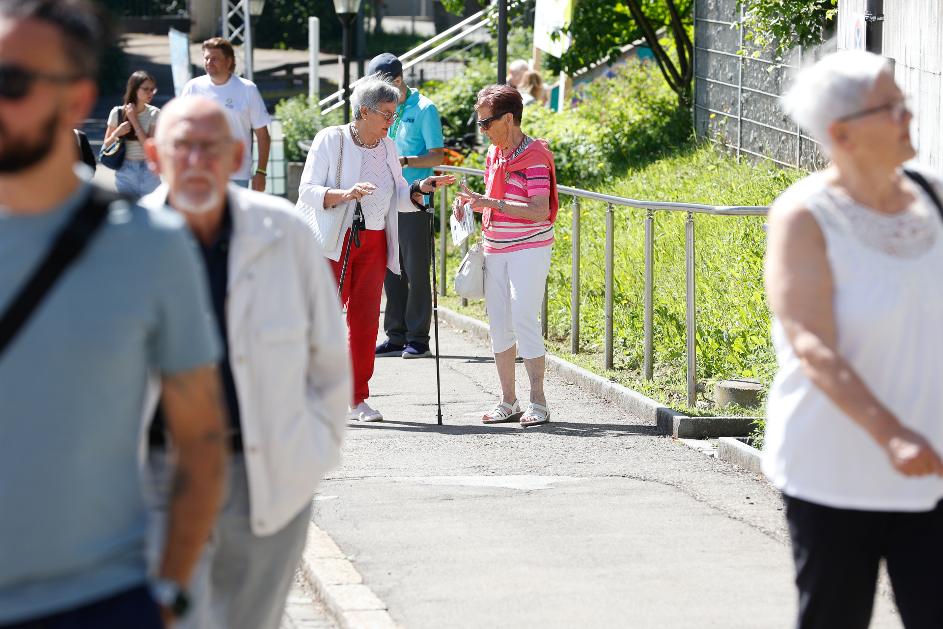An einem sonnigen Tag spazieren mehrere Menschen im Freien. In der Bildmitte unterhalten sich zwei ältere Frauen, von denen eine einen Gehstock benutzt, während im Vordergrund andere vorbeigehen, von denen einige unscharf abgebildet sind. Der Weg ist von Grünpflanzen und einem Geländer gesäumt.