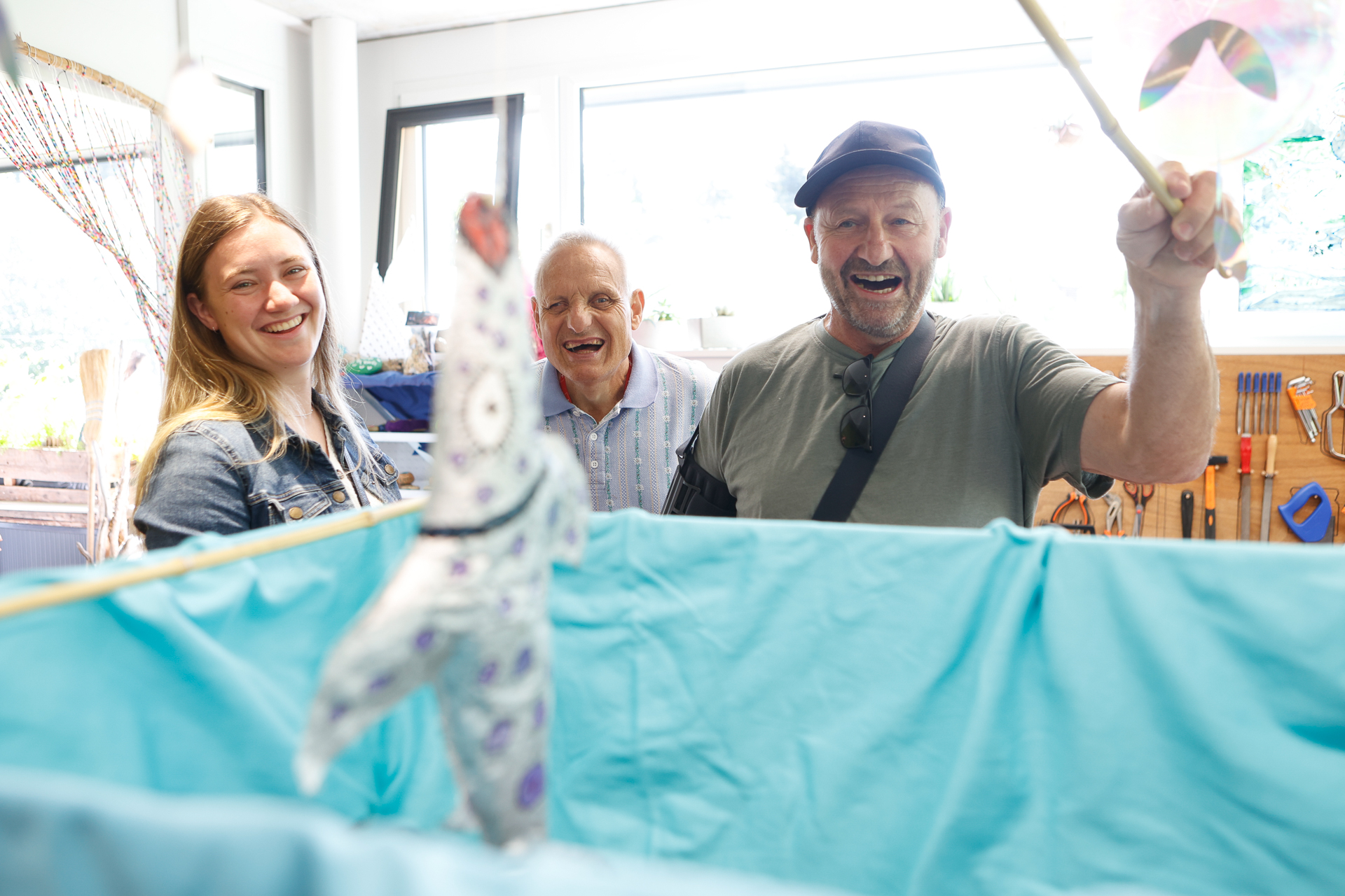 Drei Erwachsene sitzen lächelnd und lachend um eine blaue Puppenbühne herum. Eine Person hält einen Steuerhebel in der Hand und lässt eine verzierte Tierpuppe tanzen, während natürliches Licht den fröhlichen Raum erhellt.
