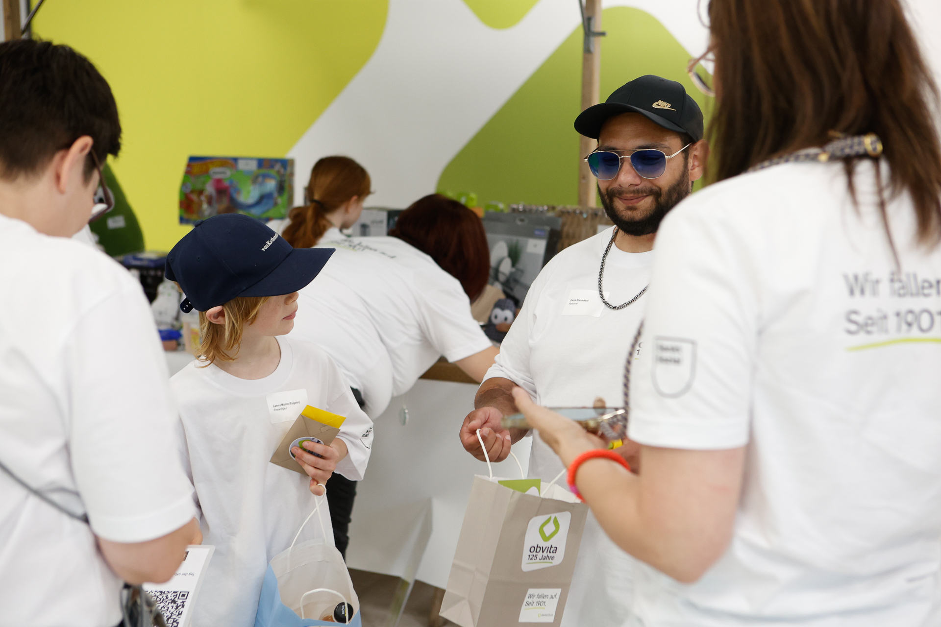 Eine Gruppe von Menschen in weißen Hemden und dunkelblauen Mützen versammelt sich in einem Raum, hält Geschenktüten in den Händen und unterhält sich. Eine Person trägt eine Sonnenbrille und eine Kette, während ein Kind lächelt; beide halten Geschenktüten mit dem „ottvita“-Logo in den Händen.