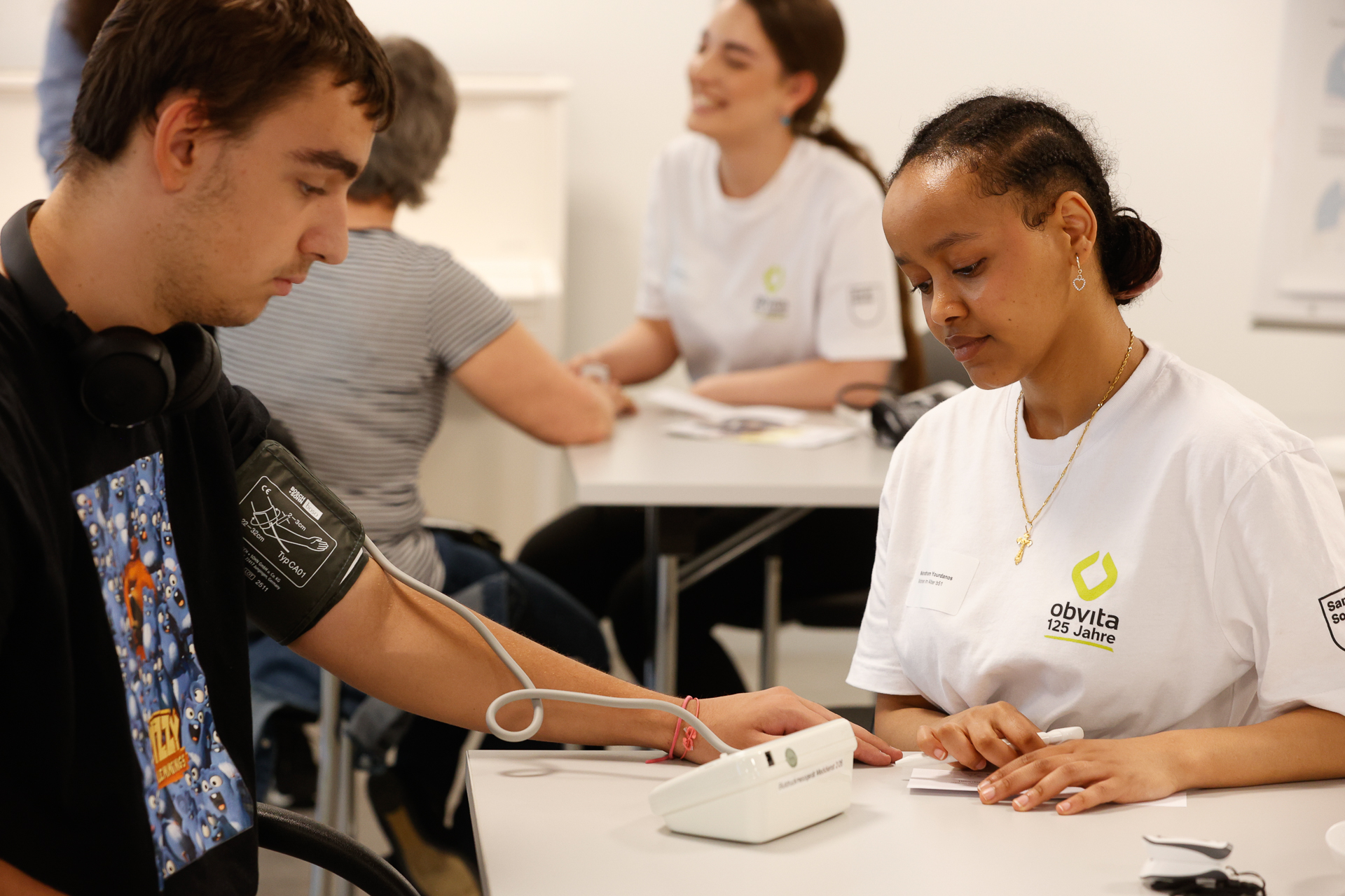Eine Frau misst an einem Tisch mit einem Blutdruckmessgerät den Blutdruck eines Mannes; beide tragen weiße Hemden. Im Hintergrund ist ein weiteres ähnliches Paar zu sehen, das ebenfalls an gesundheitsbezogenen Aktivitäten teilnimmt.
