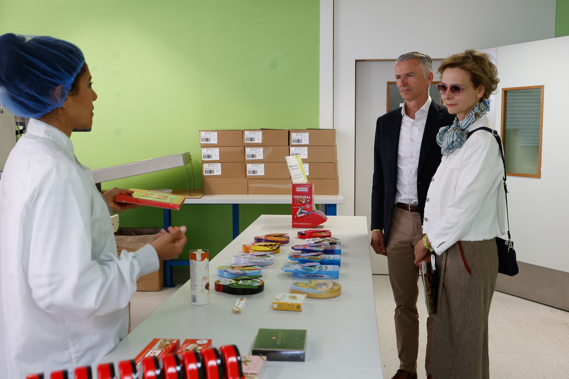 Ein Fabrikarbeiter in Laborkittel und Haarnetz präsentiert zwei professionell gekleideten Besuchern in einem hell beleuchteten Raum mit grünen Wänden und im Hintergrund aufgestapelten Kartons verschiedene verpackte Produkte, die auf einem Tisch ausliegen.