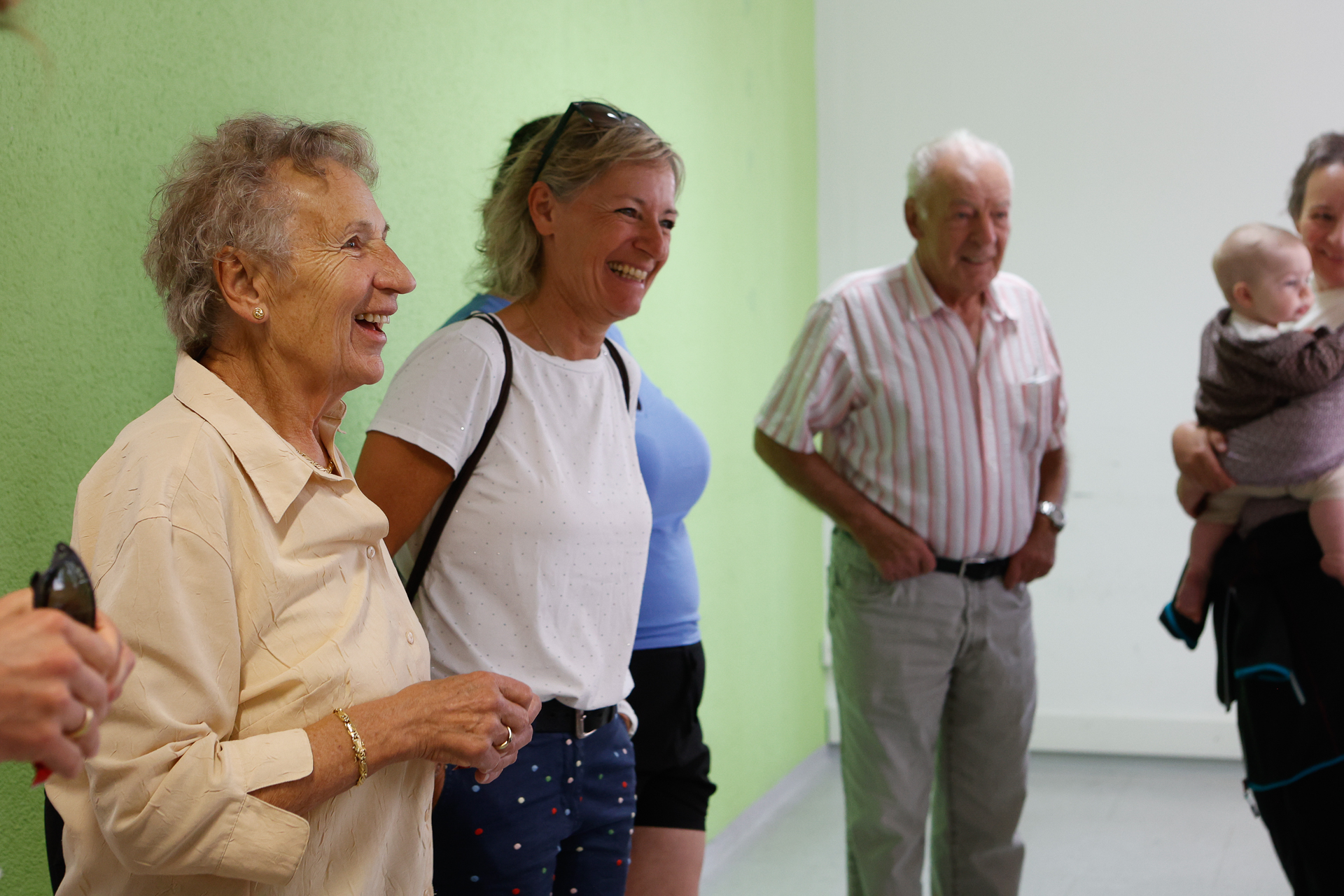 Eine Gruppe lächelnder Erwachsener, darunter zwei ältere Frauen, ein älterer Mann und eine Frau mit einem Baby im Arm, steht gemeinsam in einem Raum in der Nähe einer grünen Wand und scheint sich bei einem freundlichen Gespräch zu amüsieren.