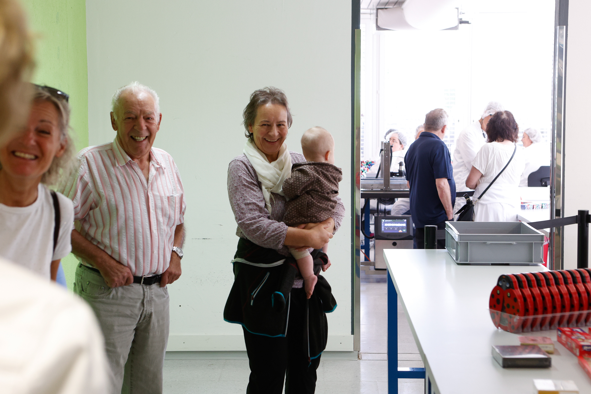 Eine Gruppe von Menschen, darunter ein lächelnder älterer Mann, eine Frau mit einem Baby im Arm und weitere Personen im Hintergrund, steht in einem hellen Raum in der Nähe einer Werkbank mit Werkzeugen und Tabletts. Alle wirken glücklich und engagiert.