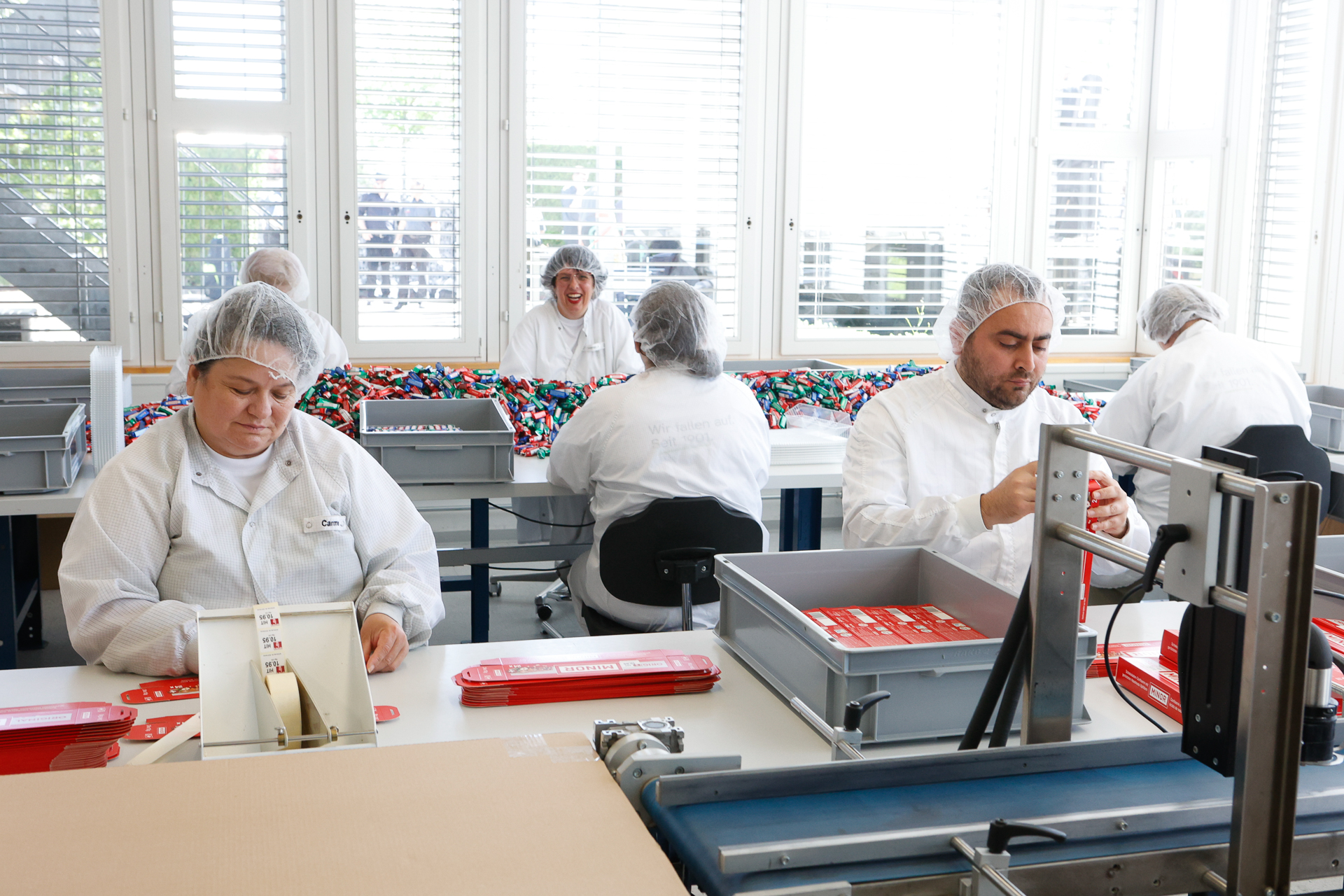 Arbeiter in weißer Schutzkleidung und Haarnetzen montieren und verpacken Artikel an einem Fließband in einer hellen, sauberen Fabrikhalle mit großen Fenstern und bunten Gegenständen im Hintergrund.