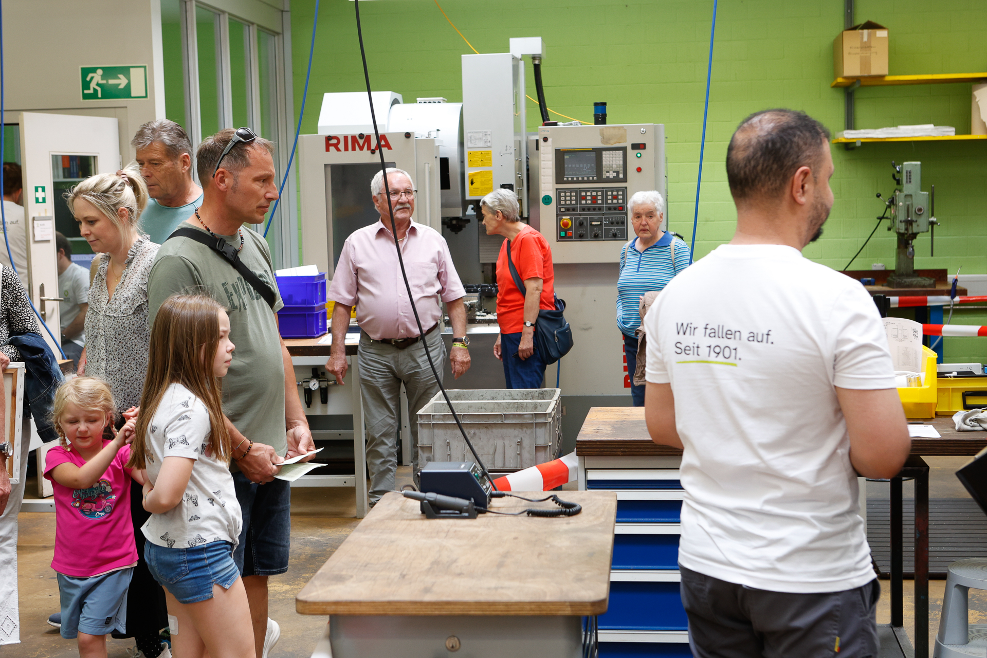Eine Gruppe von Menschen, darunter Erwachsene und Kinder, beobachtet eine Vorführung in einer Werkstatt mit Maschinen und Werkzeugen. Ein Mann im Vordergrund trägt ein weißes Hemd, auf dessen Rücken ein Schriftzug aufgedruckt ist. Der Raum hat grüne Wände.