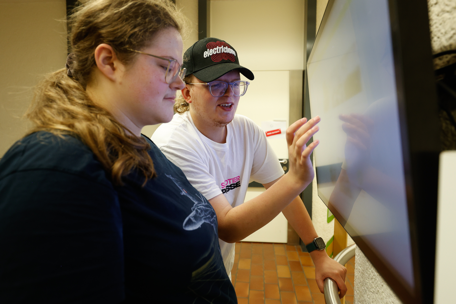 Zwei Personen stehen vor einem großen Touchscreen. Eine von ihnen zeigt auf den Bildschirm, während sie der anderen etwas erklärt. Beide wirken konzentriert und sind ganz auf das Display fixiert; die Umgebung sieht aus wie ein öffentlicher oder pädagogischer Innenraum.