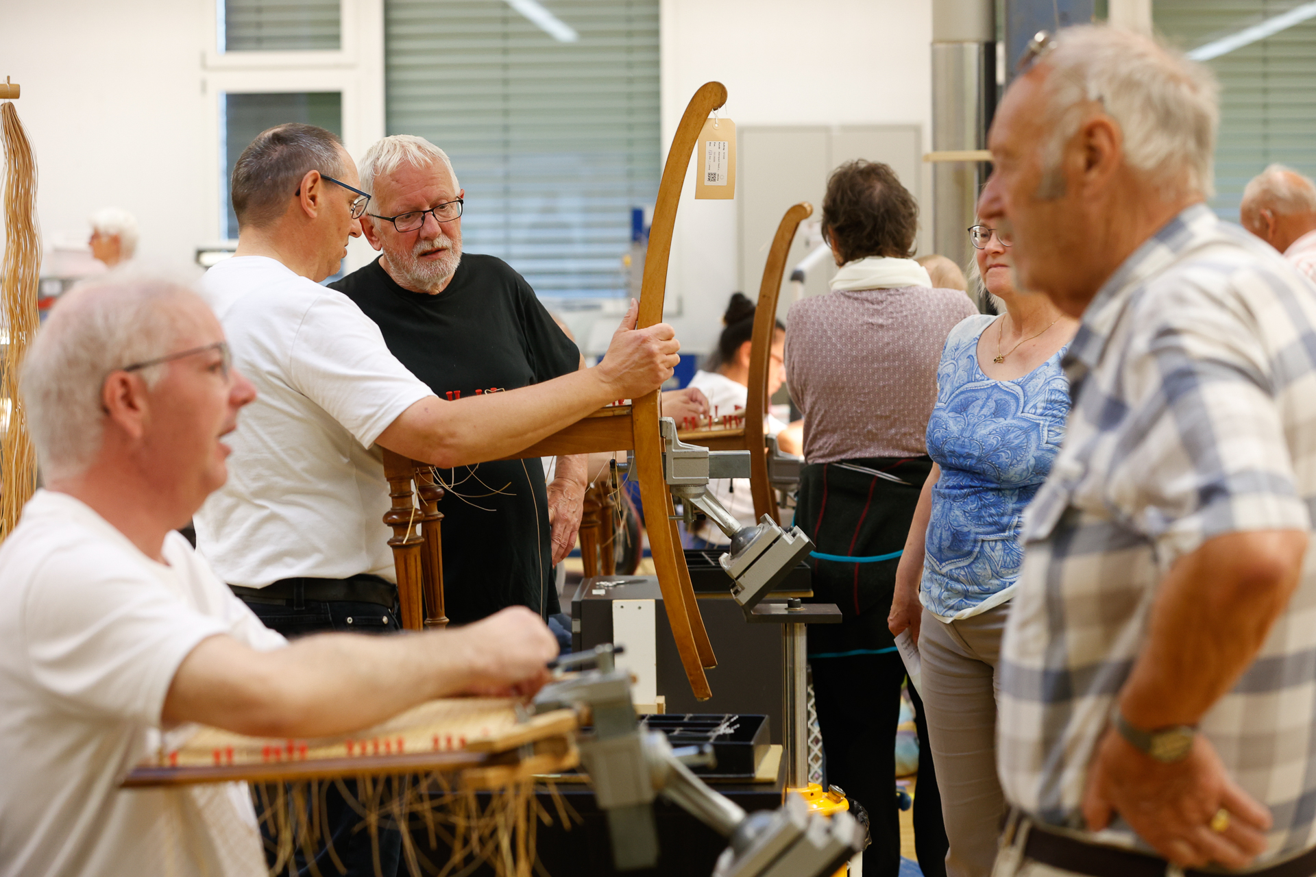 Eine Gruppe älterer Menschen steht und sitzt in einem Workshop, unterhält sich und begutachtet Gegenstände aus Holz. Werkzeuge und Webutensilien sind zu sehen, was auf eine praktische handwerkliche Tätigkeit oder eine Vorführung hindeutet.