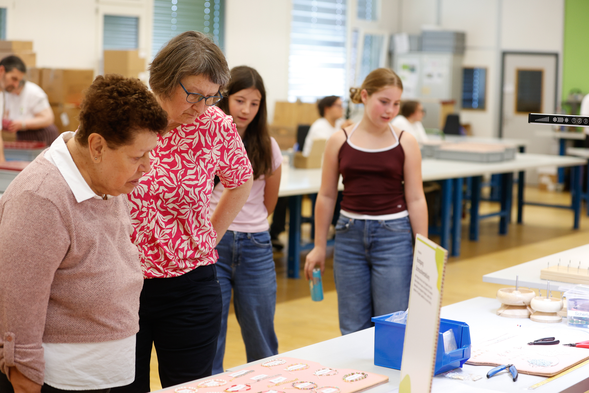 Zwei Frauen begutachten aufmerksam Gegenstände, die auf einem Tisch in einem hellen Raum ausgestellt sind, während zwei Mädchen daneben stehen und zuschauen. Im Hintergrund sind weitere Personen zu sehen, die zwischen Tischen und Kisten arbeiten.