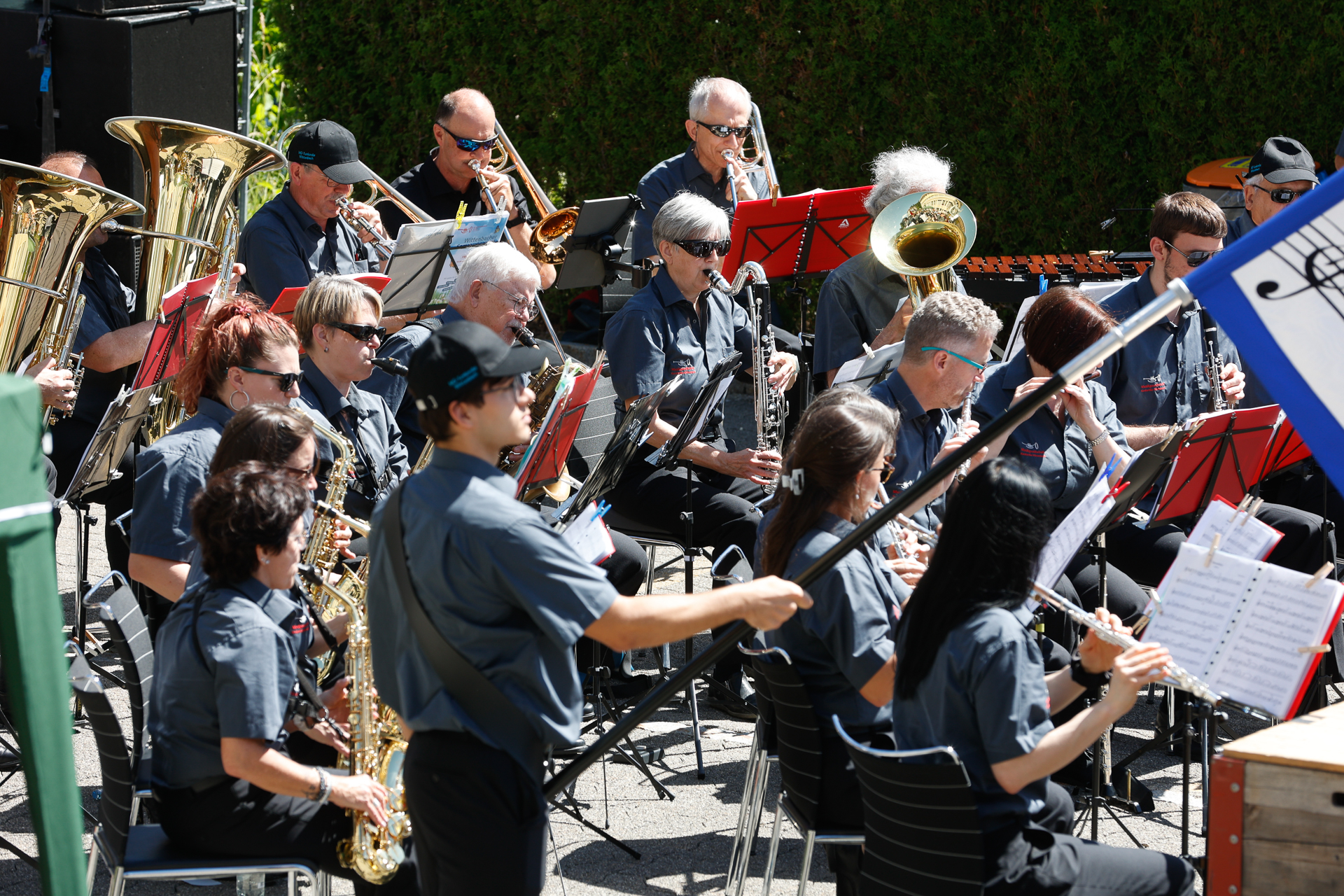 Eine große Freiluftband mit Musikern, die Blechblas-, Holzblas- und Schlaginstrumente spielen. Sie tragen einheitliche graue Hemden und Sonnenbrillen, sitzen in Reihen mit Notenständern und treten an einem sonnigen Tag auf.