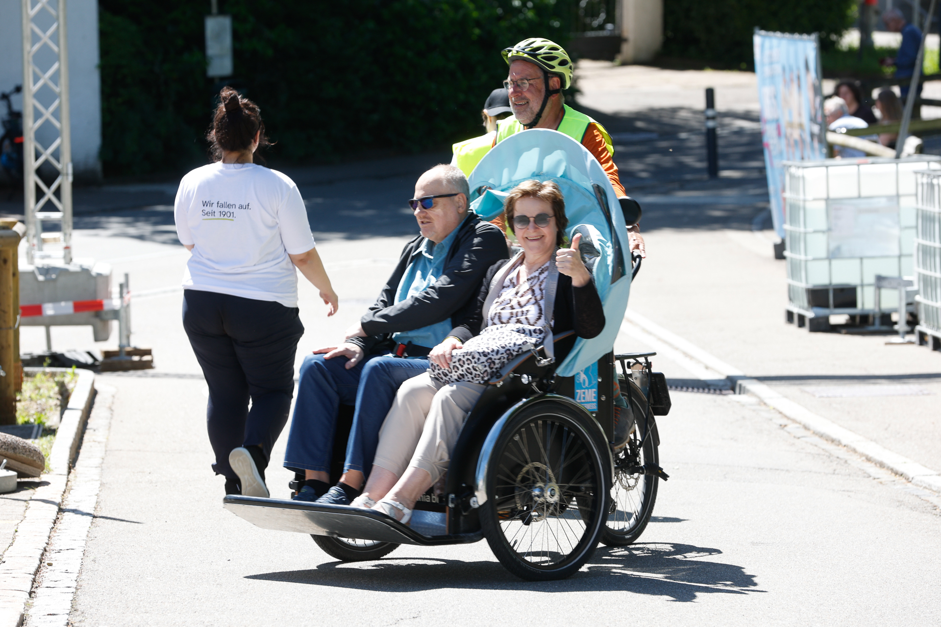 Ein Mann fährt mit einem Rikscha-Fahrrad, auf dem zwei lächelnde ältere Fahrgäste sitzen, während eine Frau vor ihnen auf einer sonnigen Straße entlanggeht. Die Fahrgäste scheinen die Fahrt zu genießen.