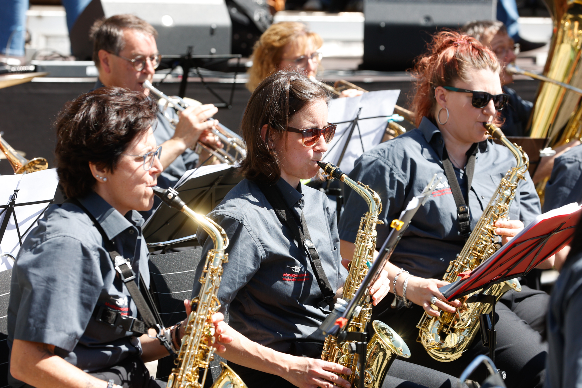 Eine Gruppe von Musikern in grauen Hemden spielt im Freien Saxophon und andere Blasinstrumente; sie sitzen in Reihen, vor sich Notenständer.